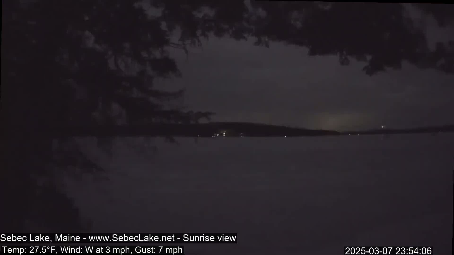 A dark, nighttime view of a calm lake with a distant shoreline, under a cloudy sky with a slight westerly wind.