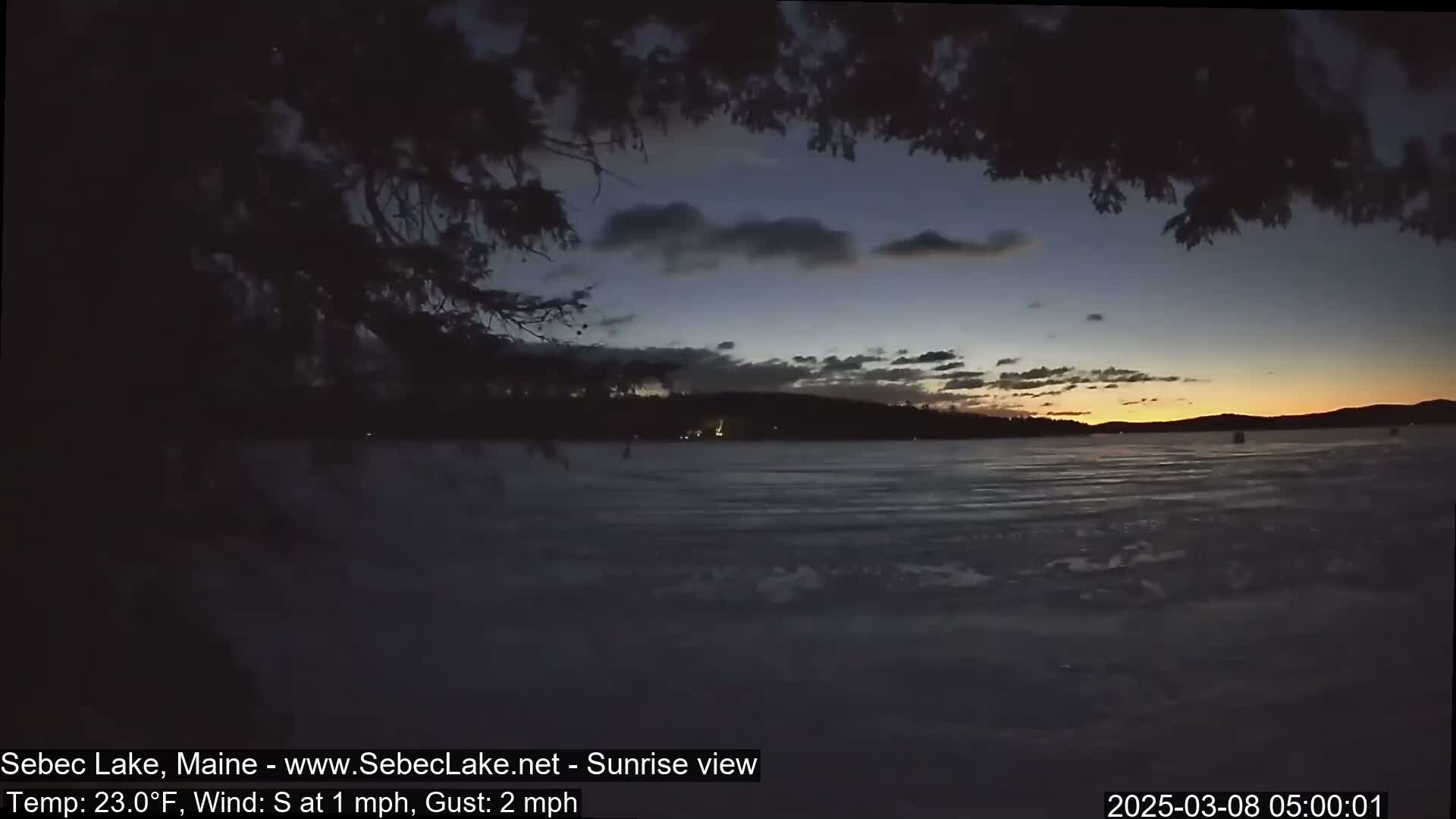A frozen lake at sunrise is viewed from behind dark tree branches, with a calm, partially cloudy sky showing orange and yellow hues on the horizon.