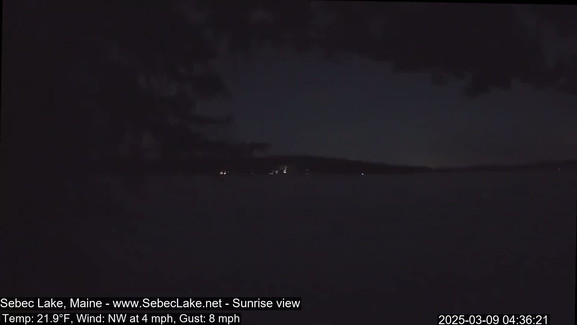 A dark, nighttime view of a lake shows a distant shoreline with faint lights under a mostly clear sky.