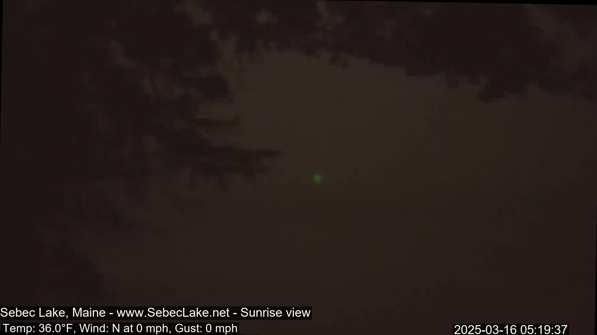 A dark, hazy view of a lake at sunrise with a calm, 36°F temperature and no wind.