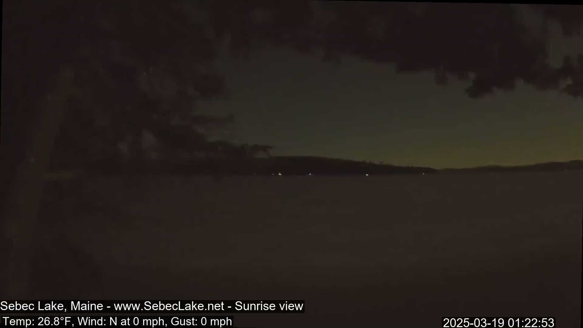 A dark, calm lake is seen at night, with a barely visible shoreline and a few faint lights in the distance under a mostly cloudy sky.