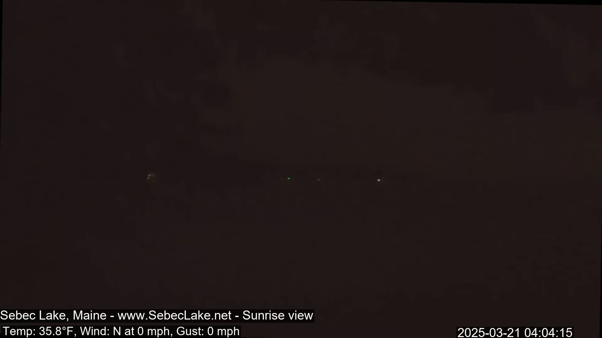 A dark, pre-dawn scene shows a few distant lights on a dark horizon under a mostly cloudy, dark sky.