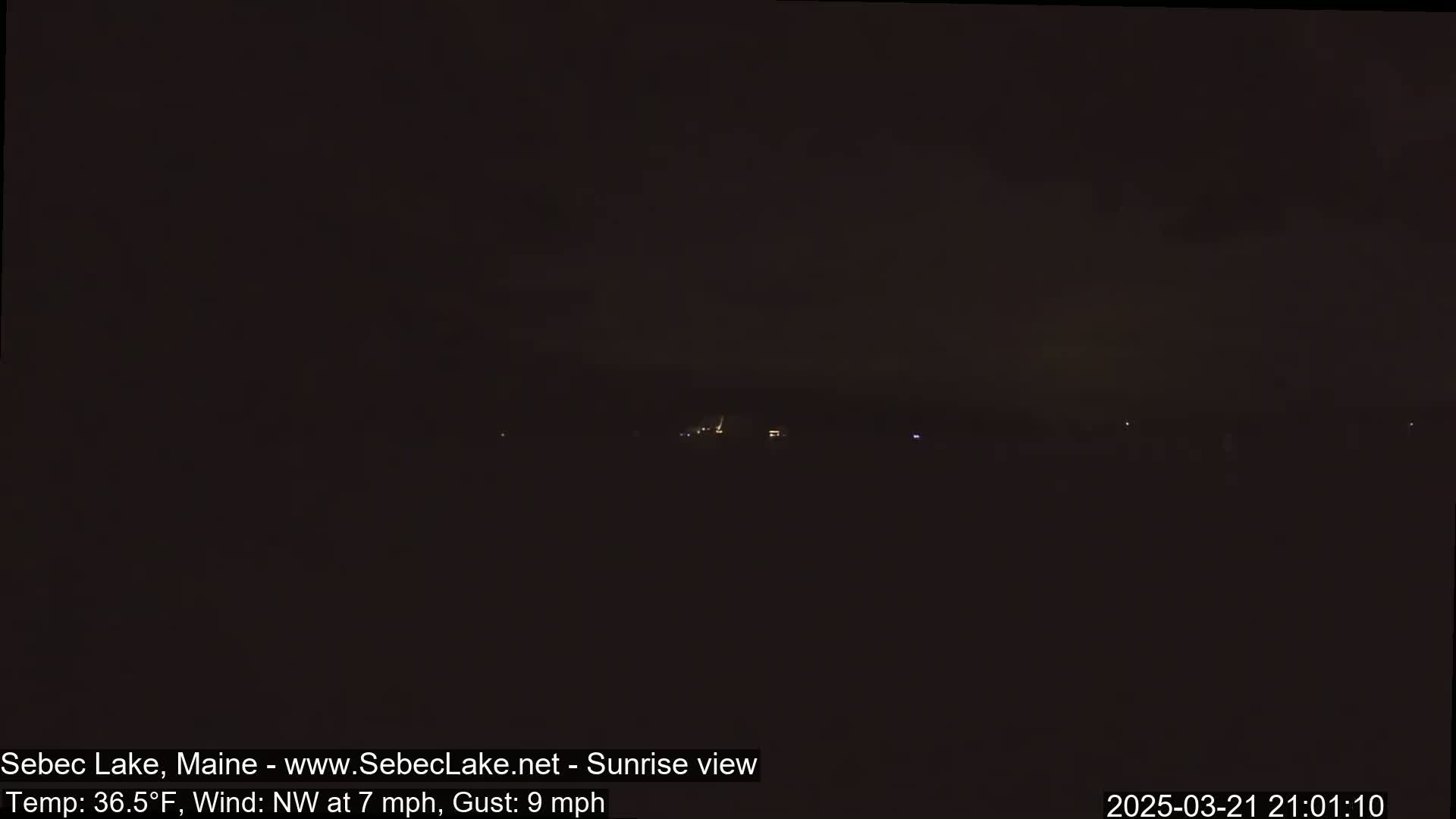 A dark, nighttime scene shows a few faint lights in the distance under a clear, dark sky with a northwest wind of 7 mph and gusts up to 9 mph.