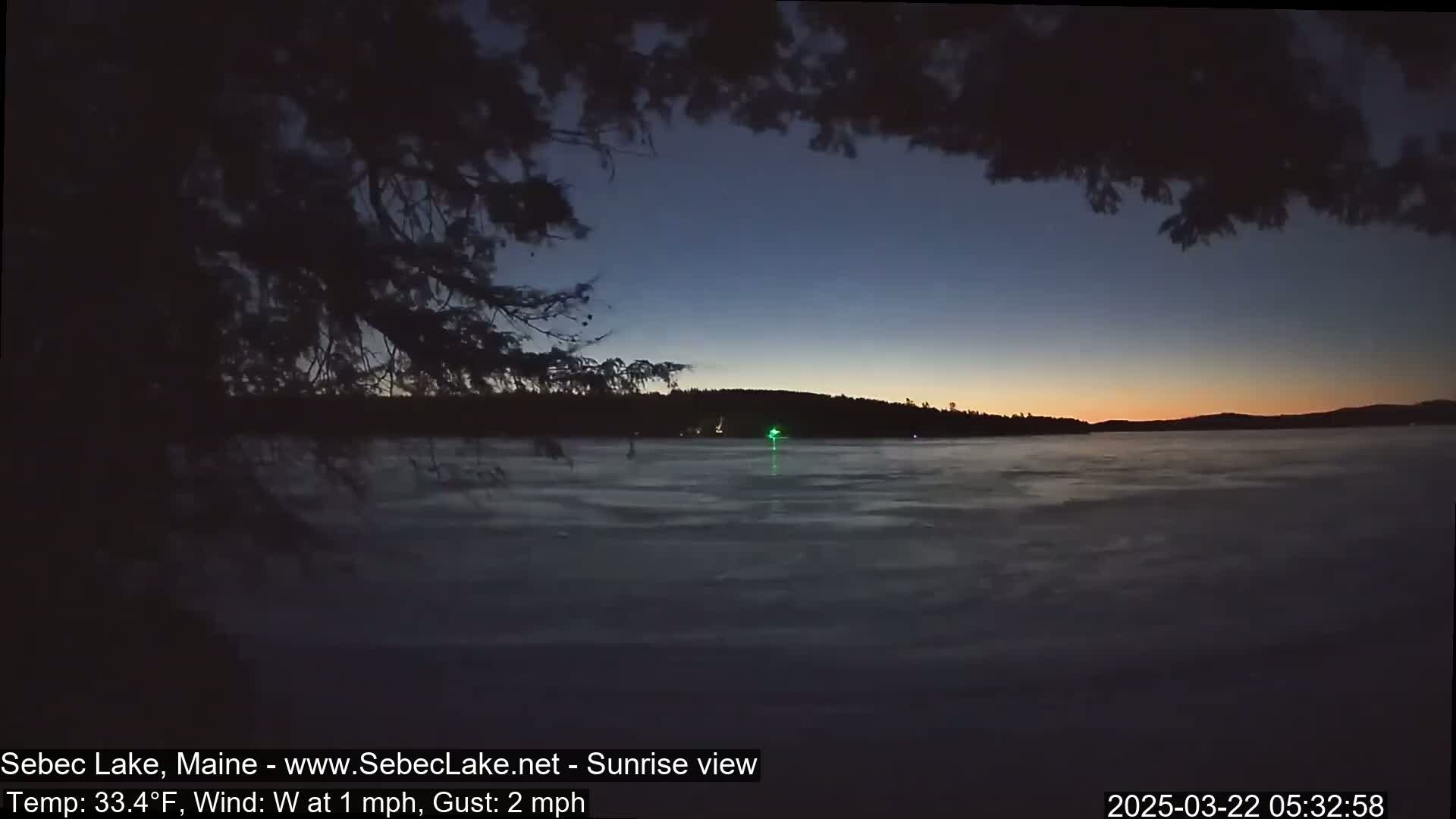 A partially frozen lake at sunrise, with a dark sky and silhouetted trees in the foreground.