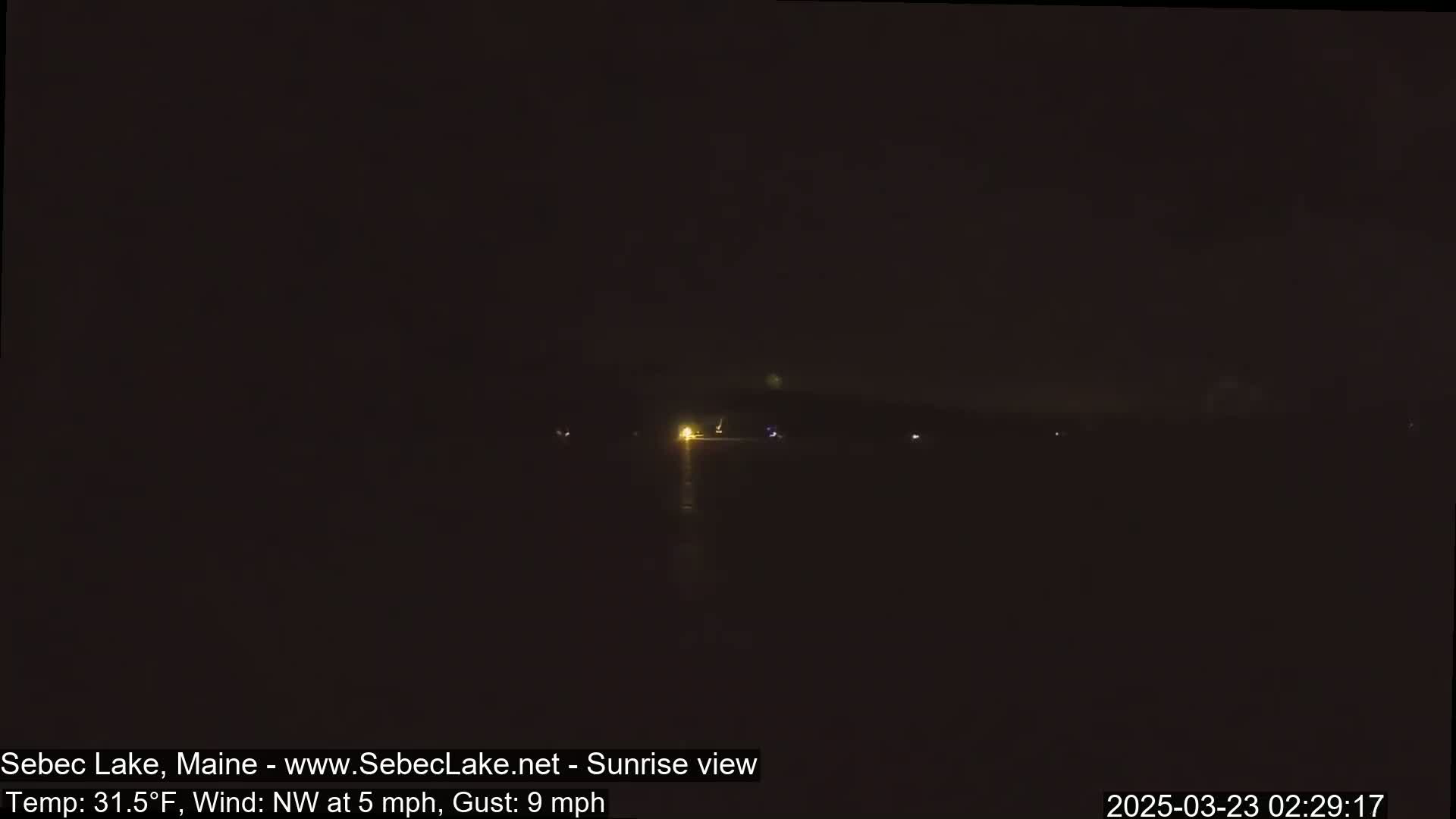A dark, nighttime view of a body of water with faint lights visible in the distance under a mostly clear sky, with a northwest wind at 5 mph and gusts up to 9 mph.