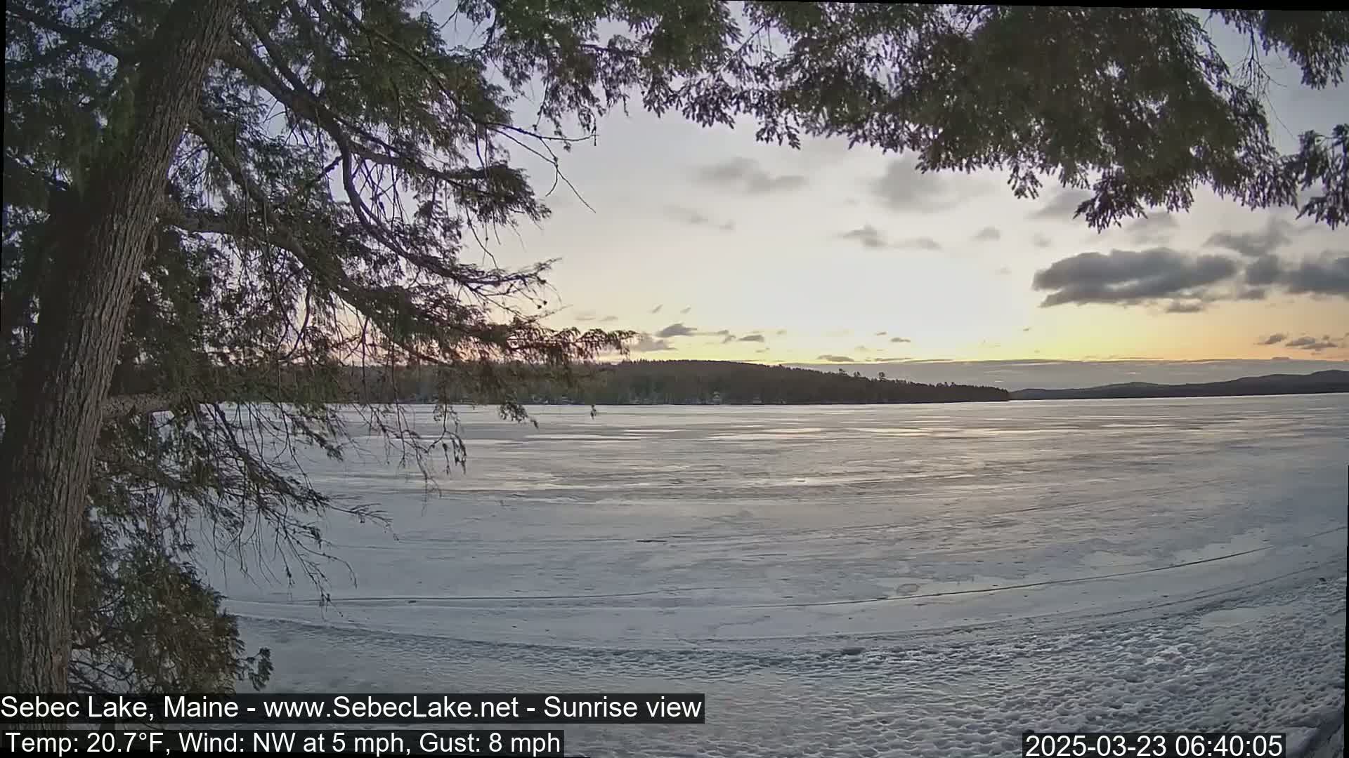 A partly cloudy sunrise over a frozen lake, viewed from behind tree branches.