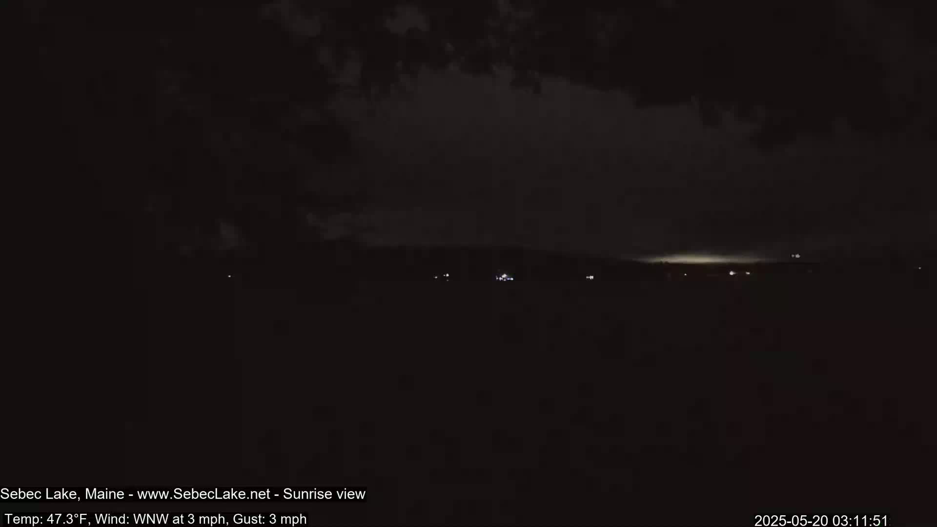 A dark, nighttime landscape shows a faint, light horizon with a few scattered lights in the distance under a cloudy sky.