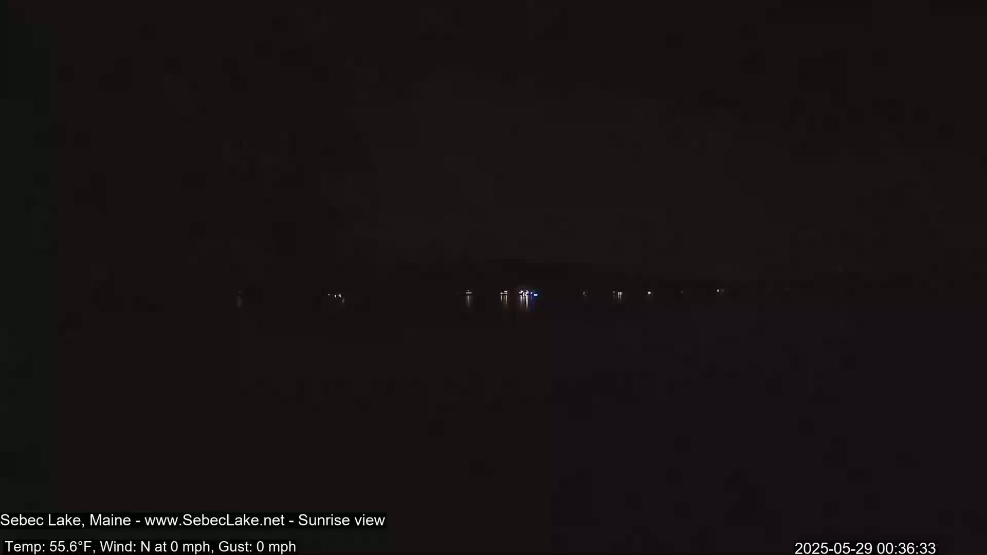 A nighttime view of a dark body of water with a few faint lights visible on the far shore under a dark sky.