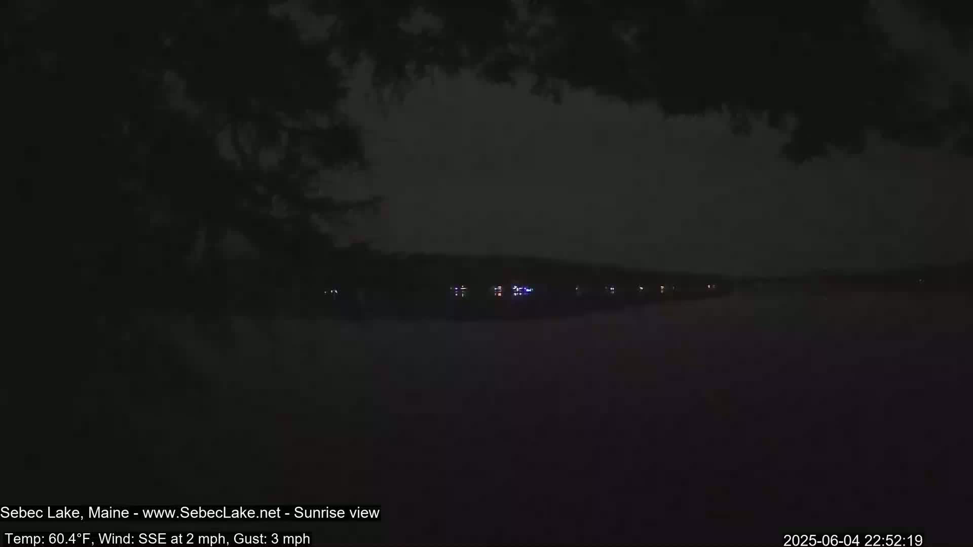 A dark, nighttime view of a lake with a few distant lights across the water under a mostly cloudy sky.
