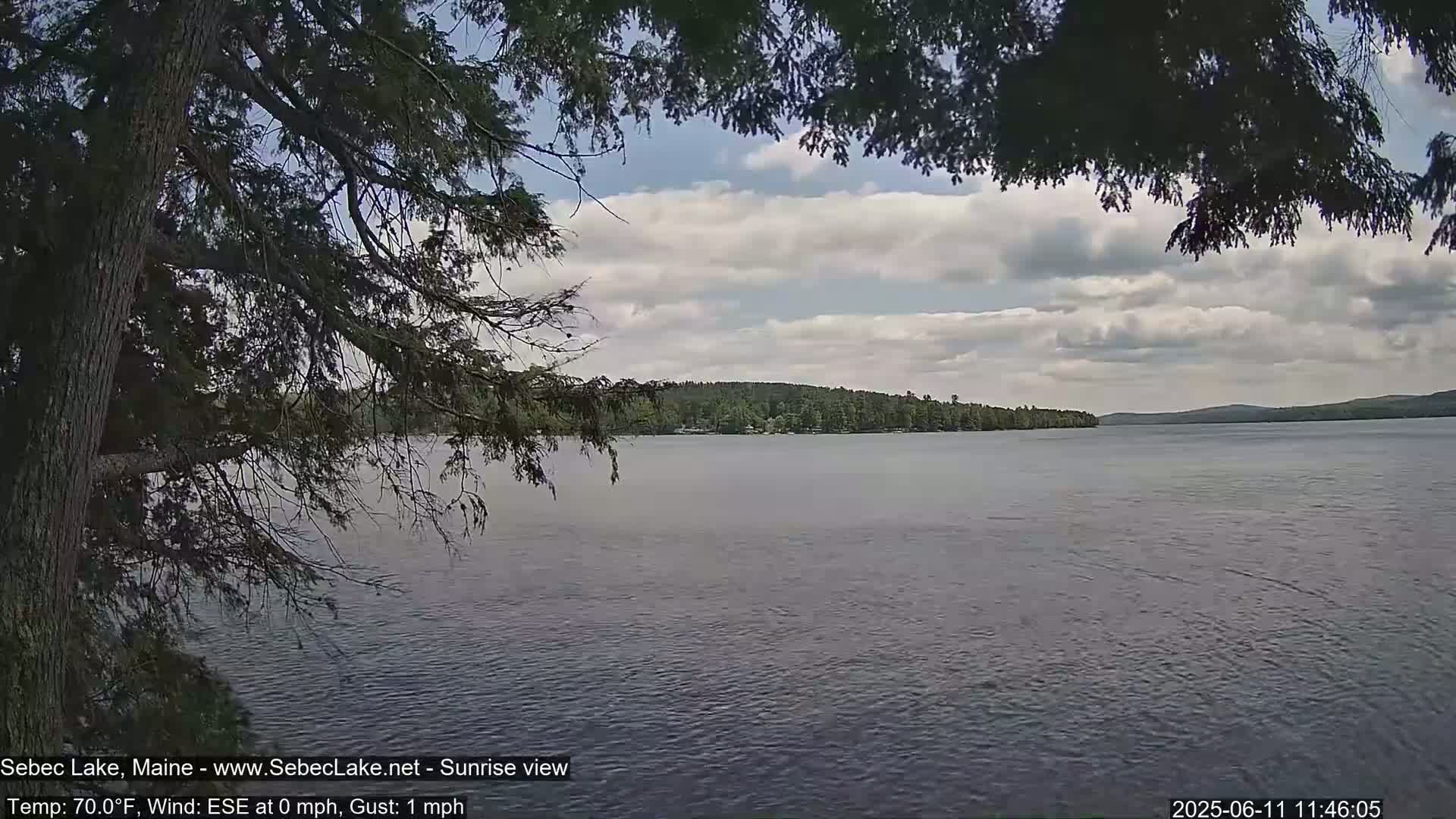 A partly cloudy day shows a view of a calm lake with a tree-lined shore and forested hills in the distance.