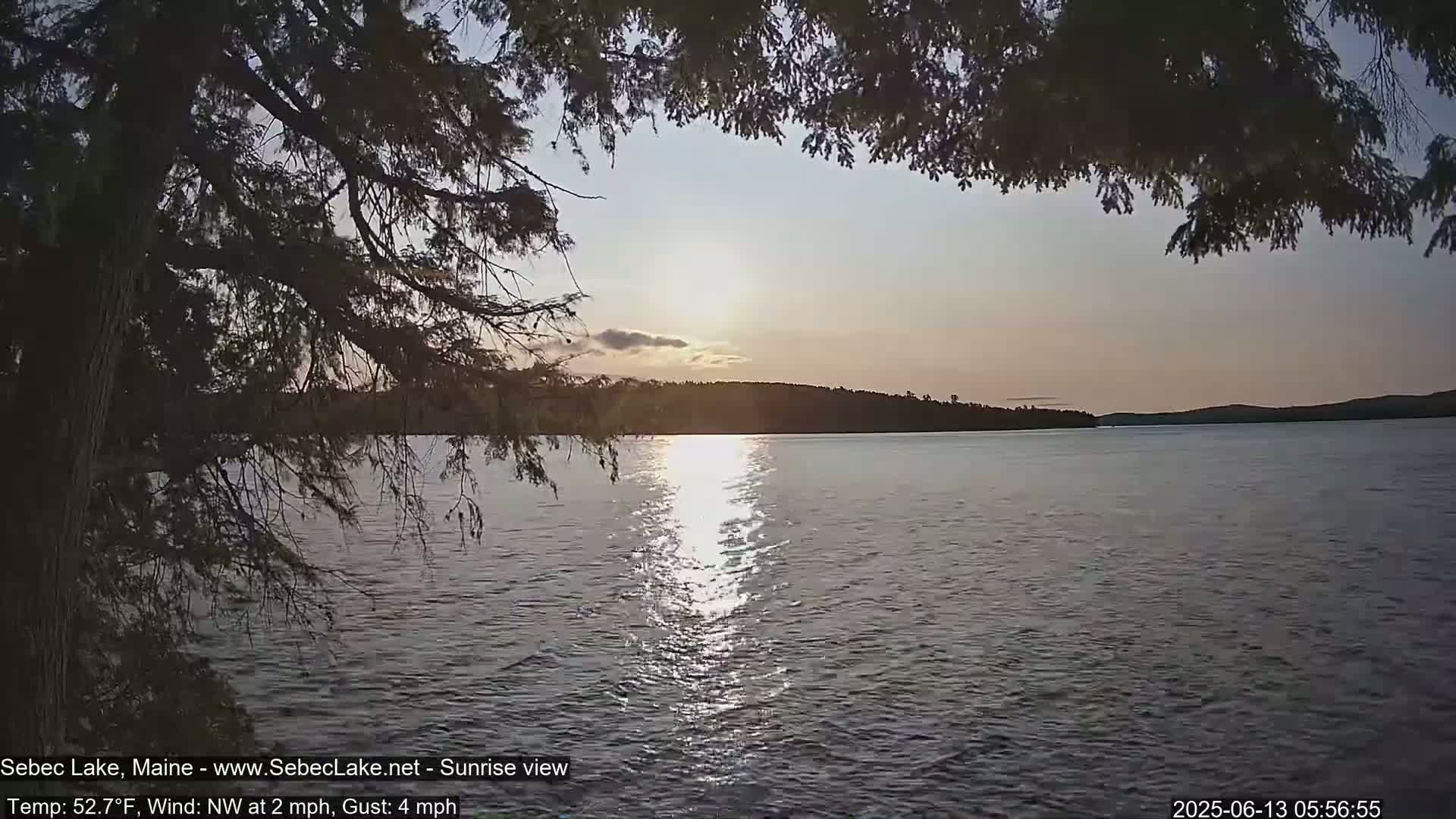 A calm lake at sunrise, partially viewed through overhanging tree branches, with a gentle northwest wind.
