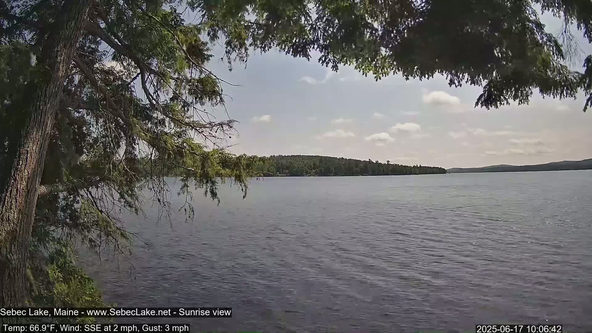 A partly cloudy sky is seen over a calm lake with a tree's branches partially obscuring the view from the foreground.