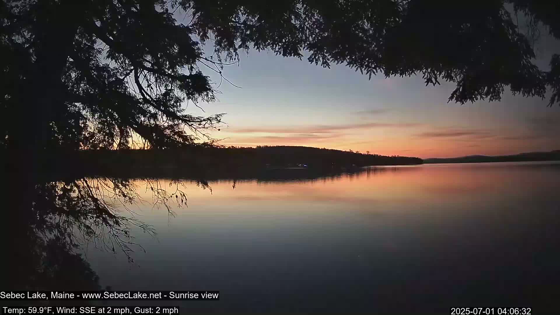 A tranquil lake at sunrise, viewed from under the branches of a silhouetted tree, with calm waters reflecting the pastel sky.
