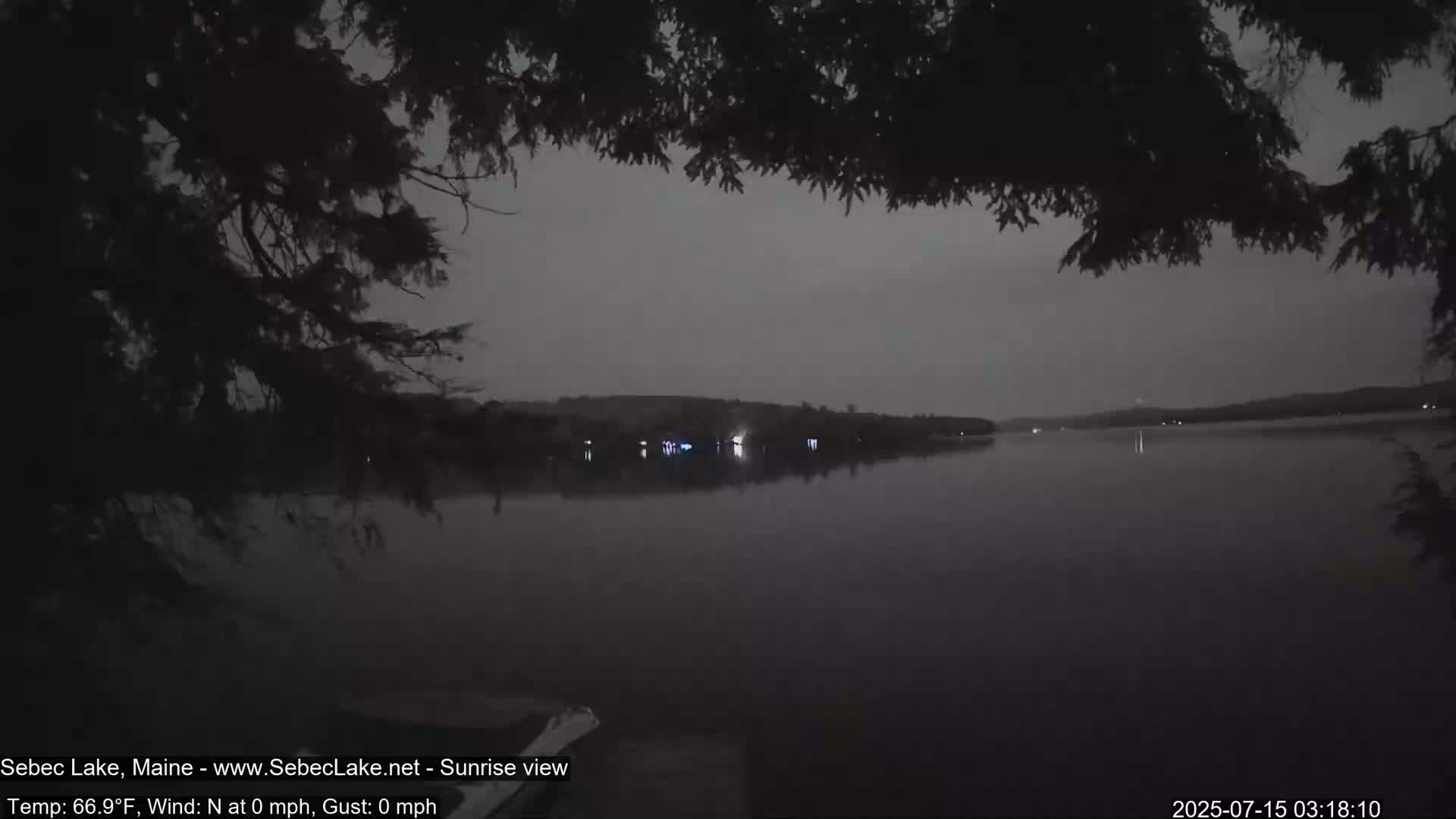 A calm, dark lake is seen at twilight, viewed from behind tree branches.