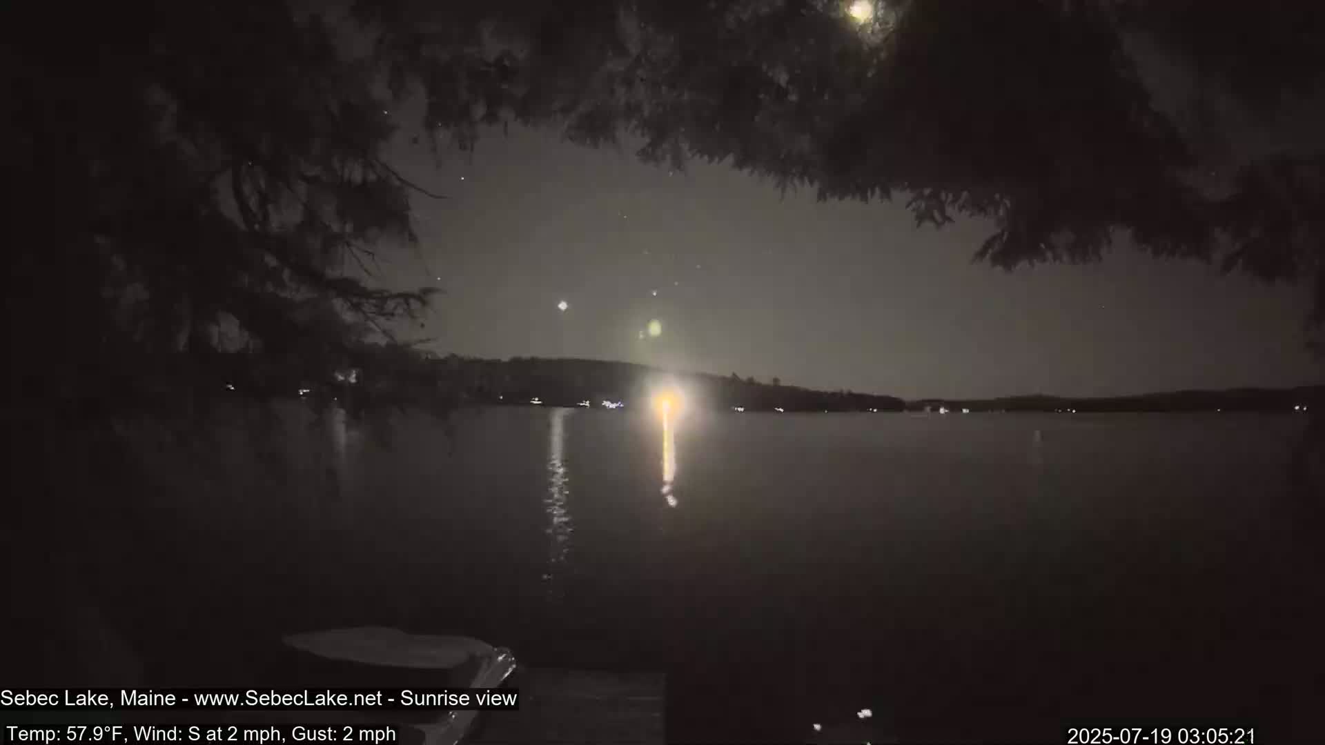 A nighttime view of a calm lake under a partly cloudy sky, with faint lights visible across the water and tree branches partially obscuring the view from a lakeside vantage point.
