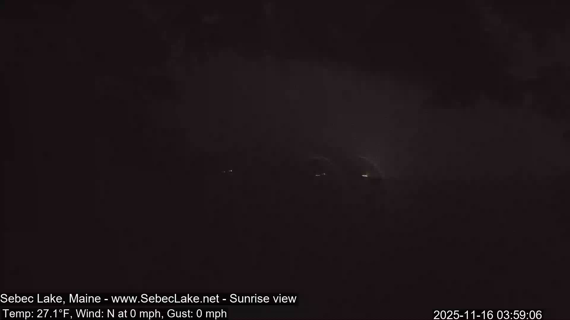 An extremely dark, pre-dawn or nighttime outdoor scene depicts a few tiny, distant lights against a backdrop of what appears to be an overcast sky.