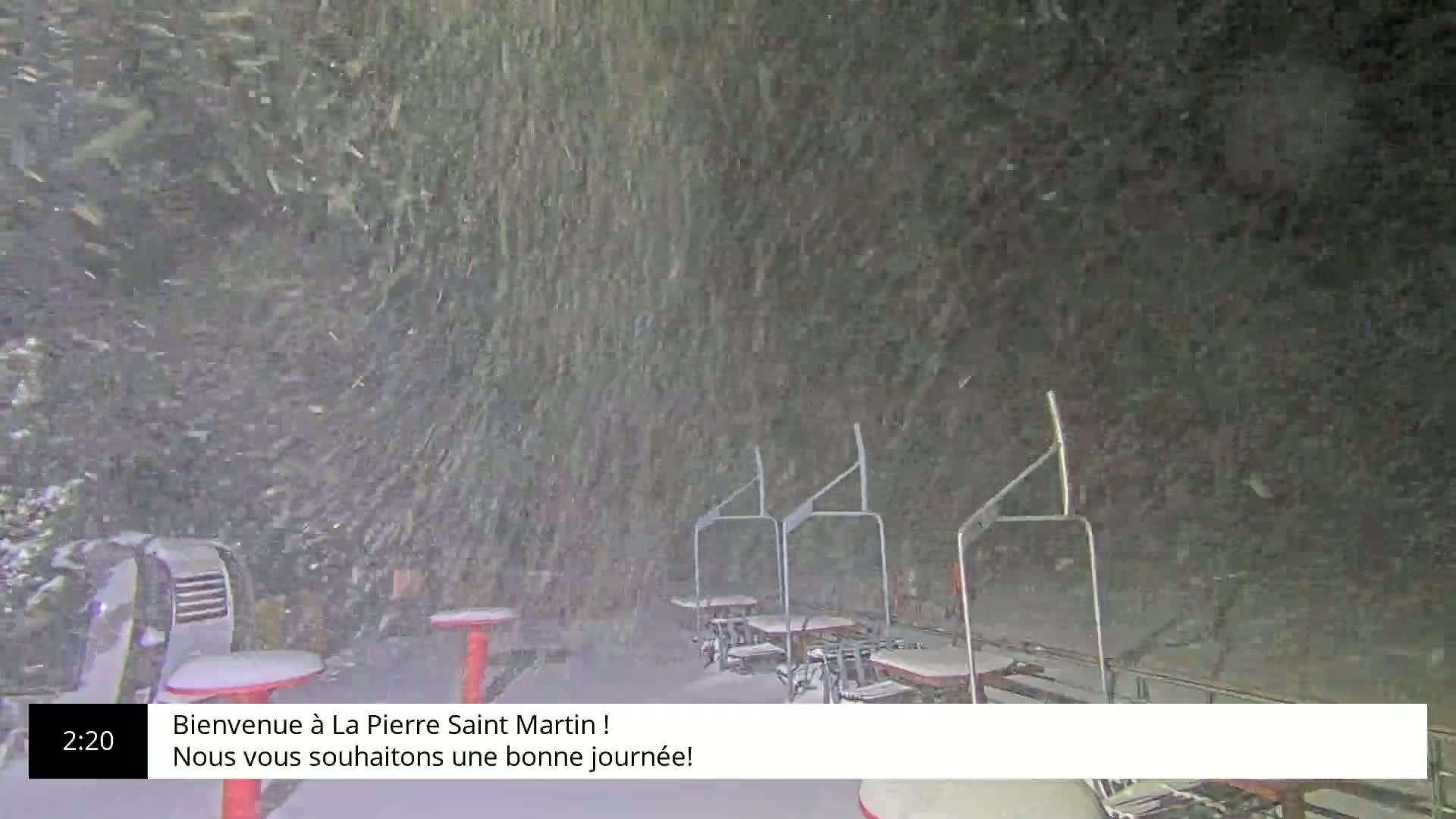 A snowstorm blankets a ski lift area with several empty chairlifts and small tables covered in snow.