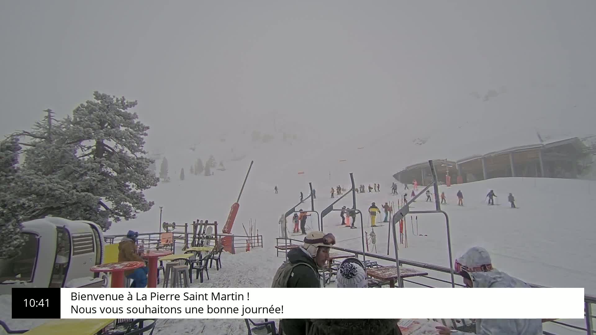 A snowy ski slope with several skiers and snowboarders visible in the hazy, overcast weather, alongside an outdoor seating area with people gathered around tables.