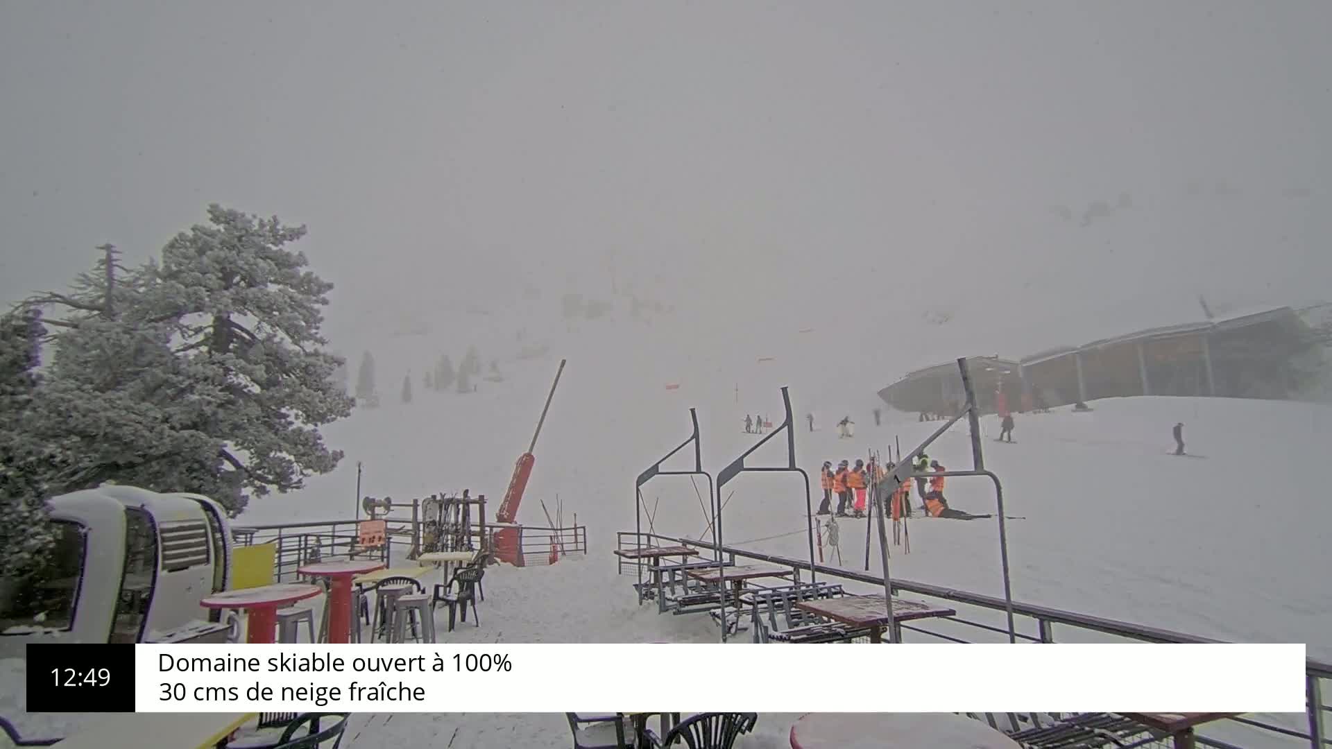 A snow-covered ski resort is seen during a heavy snowstorm, with several skiers and a lift visible in the distance.