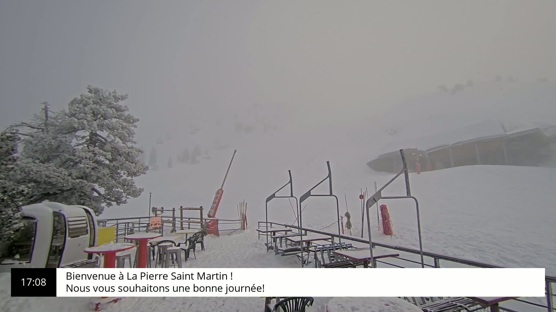 A snow-covered outdoor patio area with several tables and chairs is visible through heavy fog, next to a ski lift.
