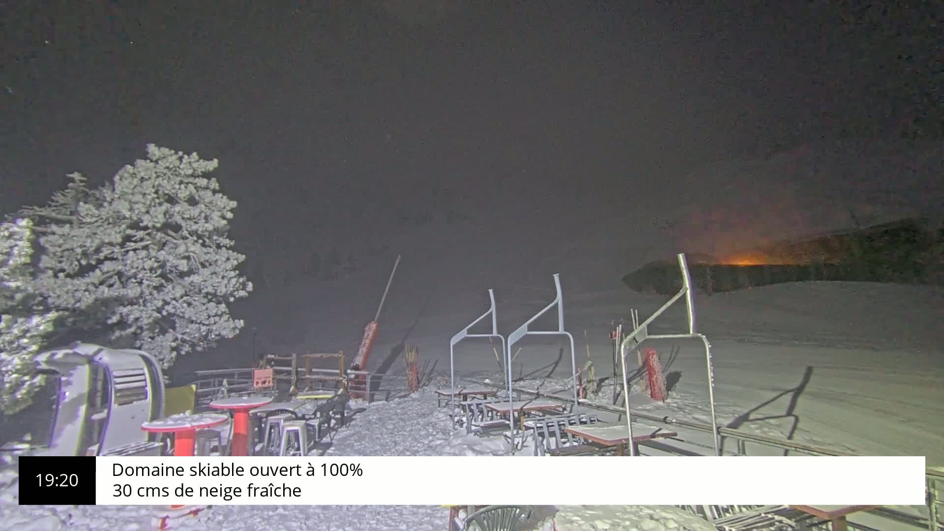 A snow-covered outdoor ski resort area at night, under a dark, cloudy sky, shows several empty tables and chairs near a ski lift.