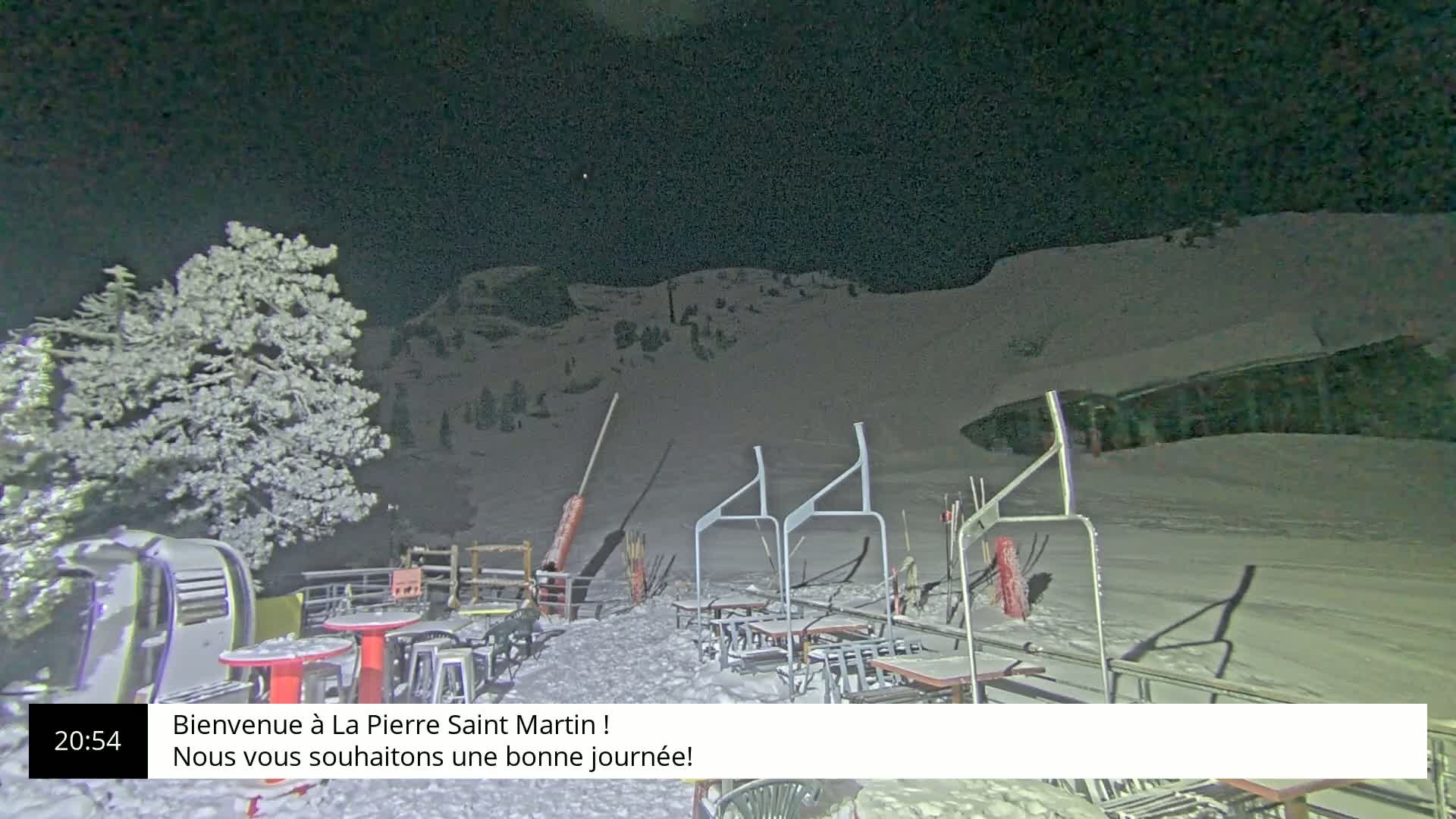 A snow-covered outdoor patio with tables and chairs sits under a dark, clear night sky next to a ski lift and snowy hill.