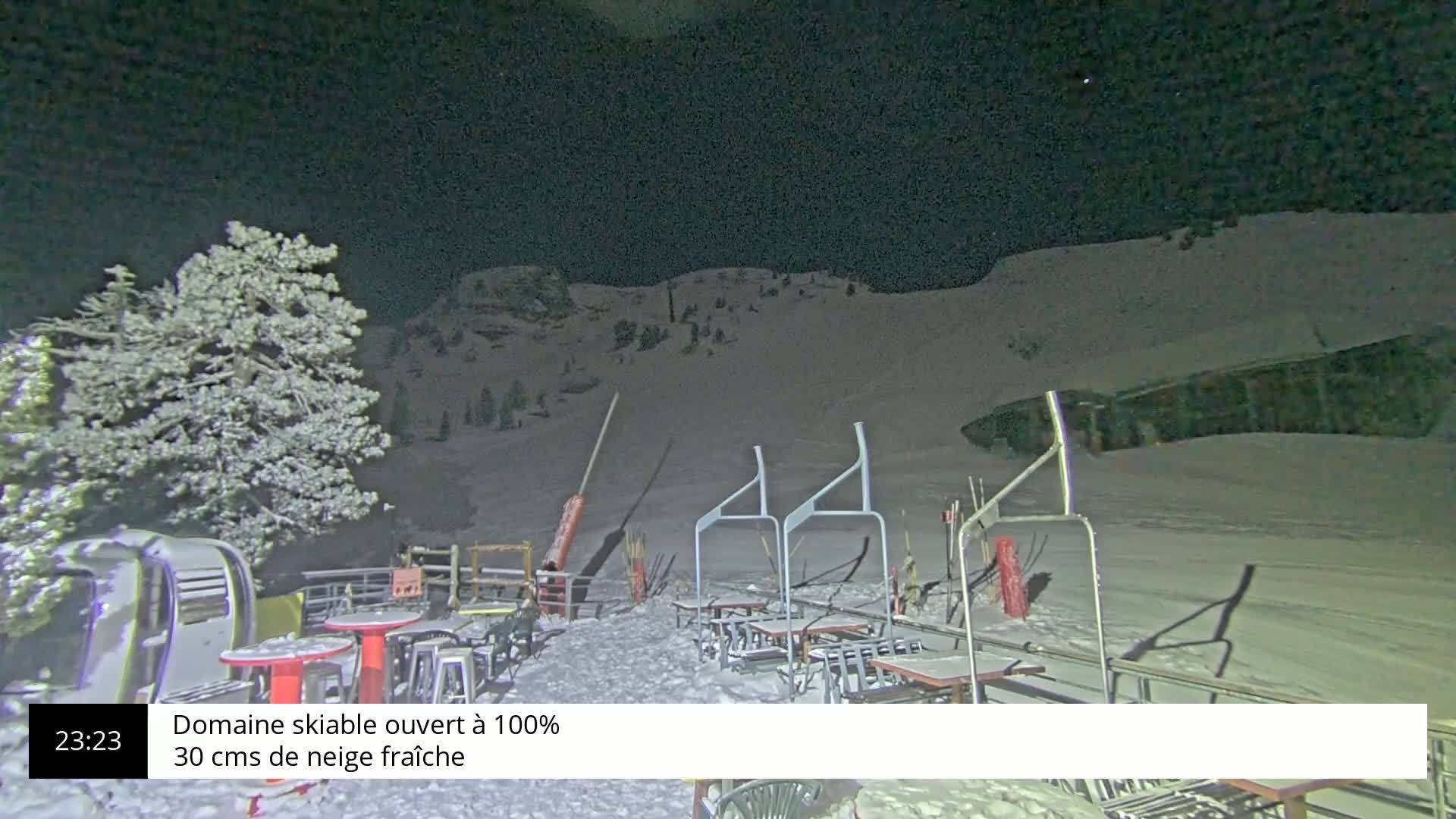 A snow-covered ski slope at night, with outdoor seating and a snow-laden tree in the foreground.