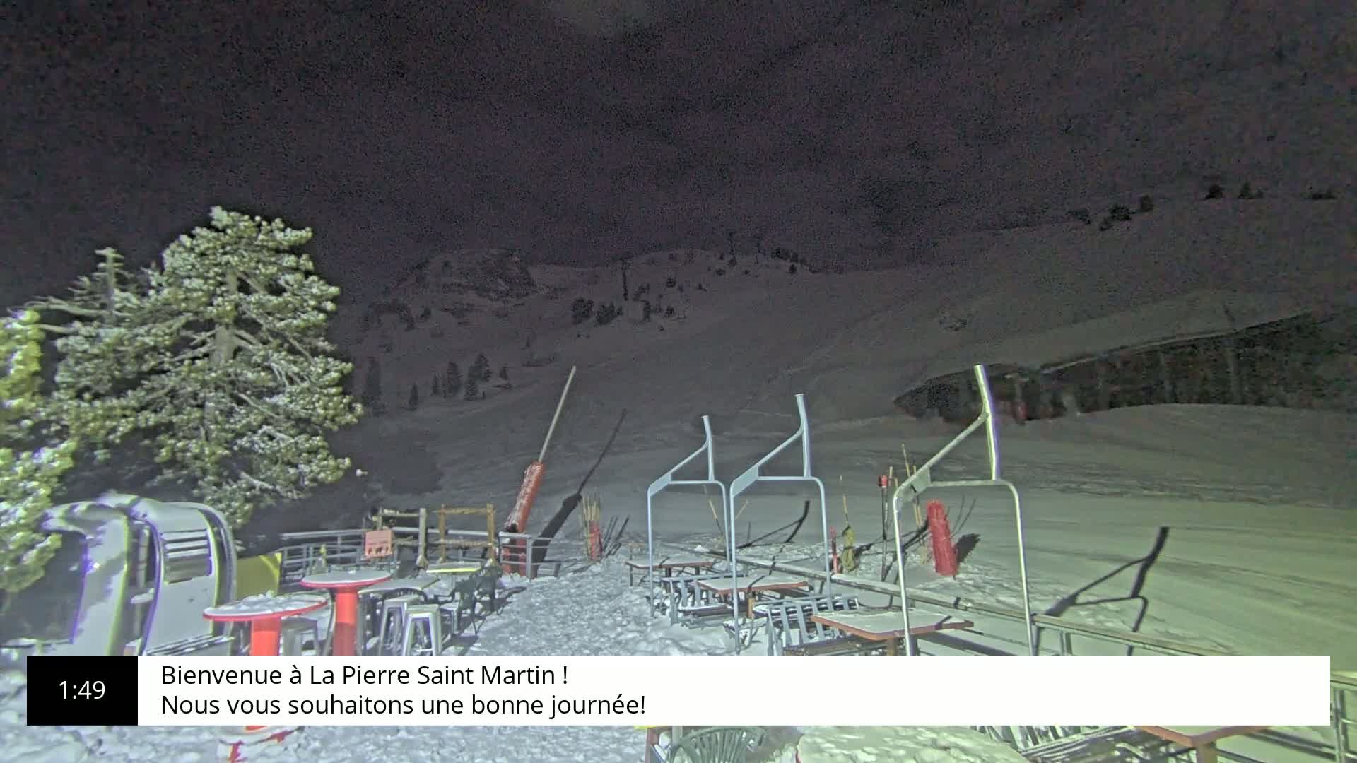 A snow-covered ski slope at night, illuminated by artificial light, with outdoor seating and a chairlift visible.