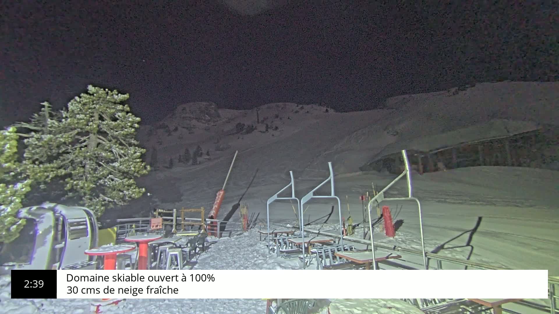 A nighttime view of a snow-covered ski slope with outdoor seating and a snow-laden tree in the foreground under a dark sky.