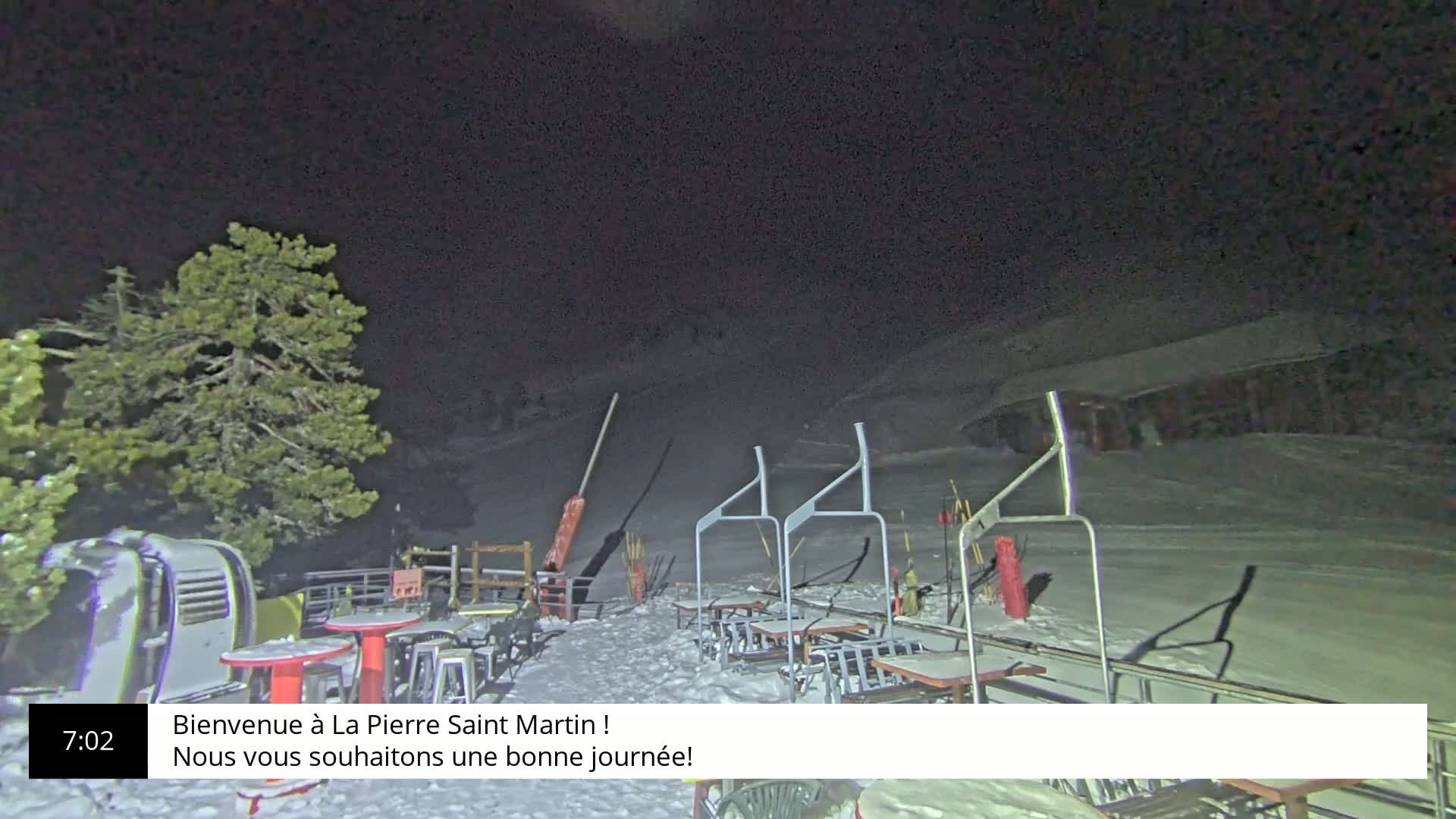A snow-covered outdoor seating area with tables and chairs sits under a dark night sky next to a ski lift.