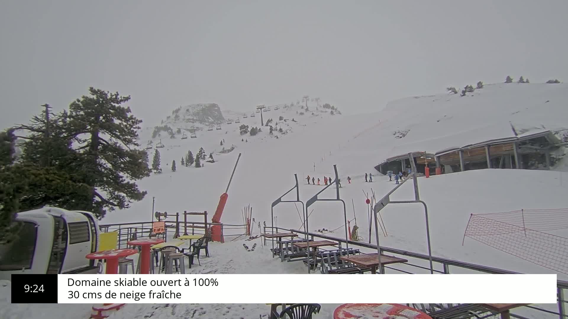 A snowy ski resort on an overcast day shows several skiers on the slopes, chairlifts, and outdoor seating at a restaurant.