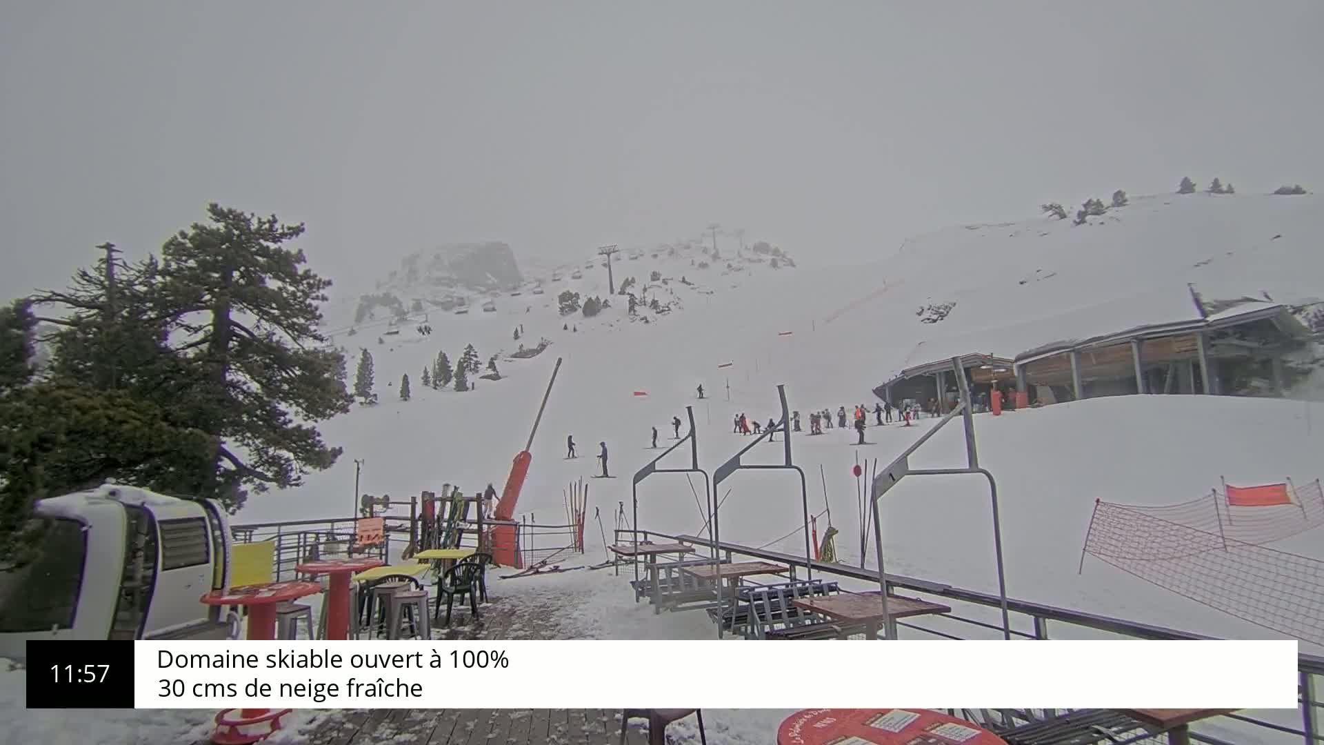 A snowy ski slope with several skiers and a ski lift, viewed from a patio with tables and chairs, under an overcast sky.