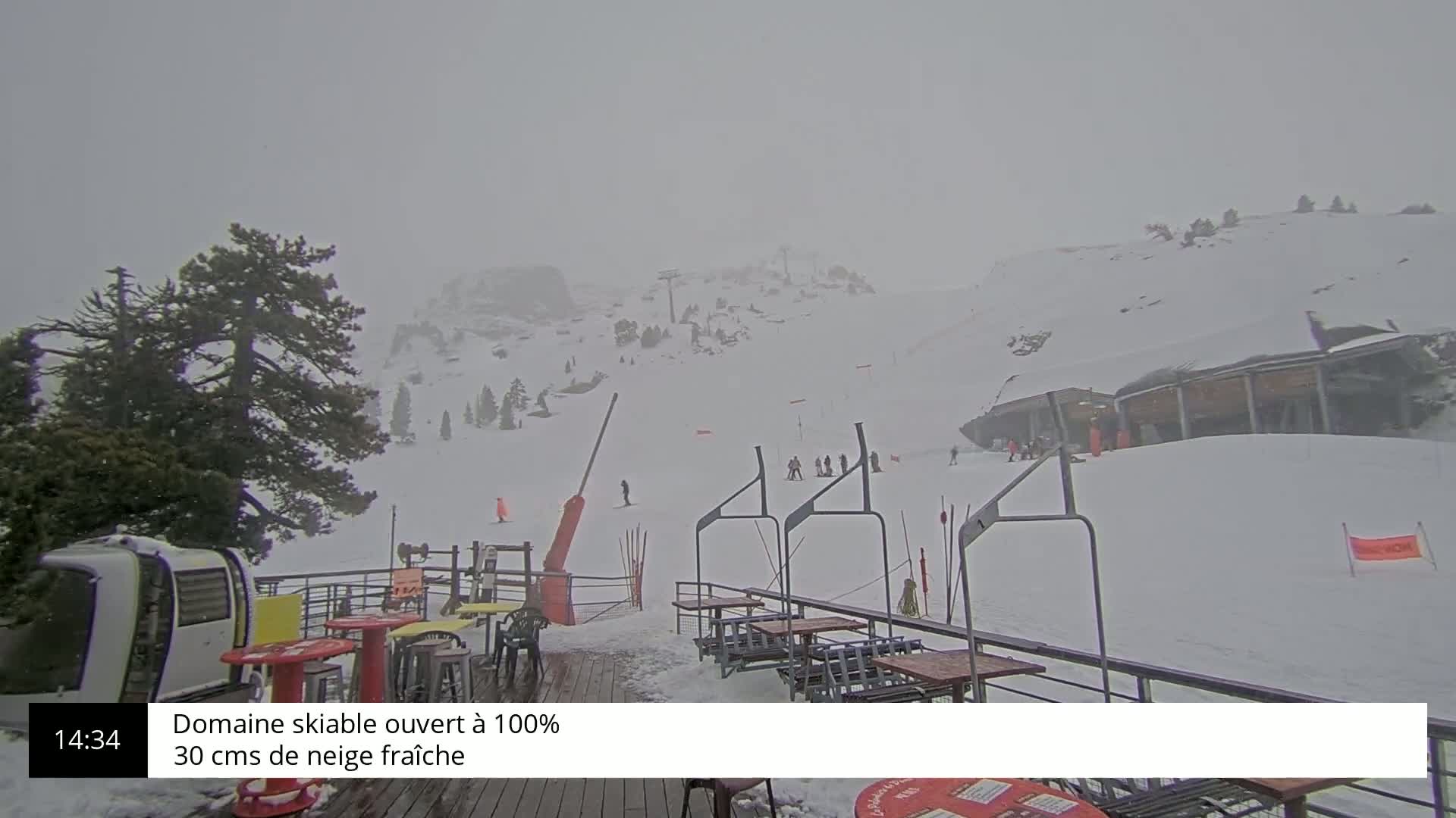 A snowy ski slope with a few skiers visible in the distance, under hazy, overcast conditions, is viewed from an outdoor patio area with tables and chairs.