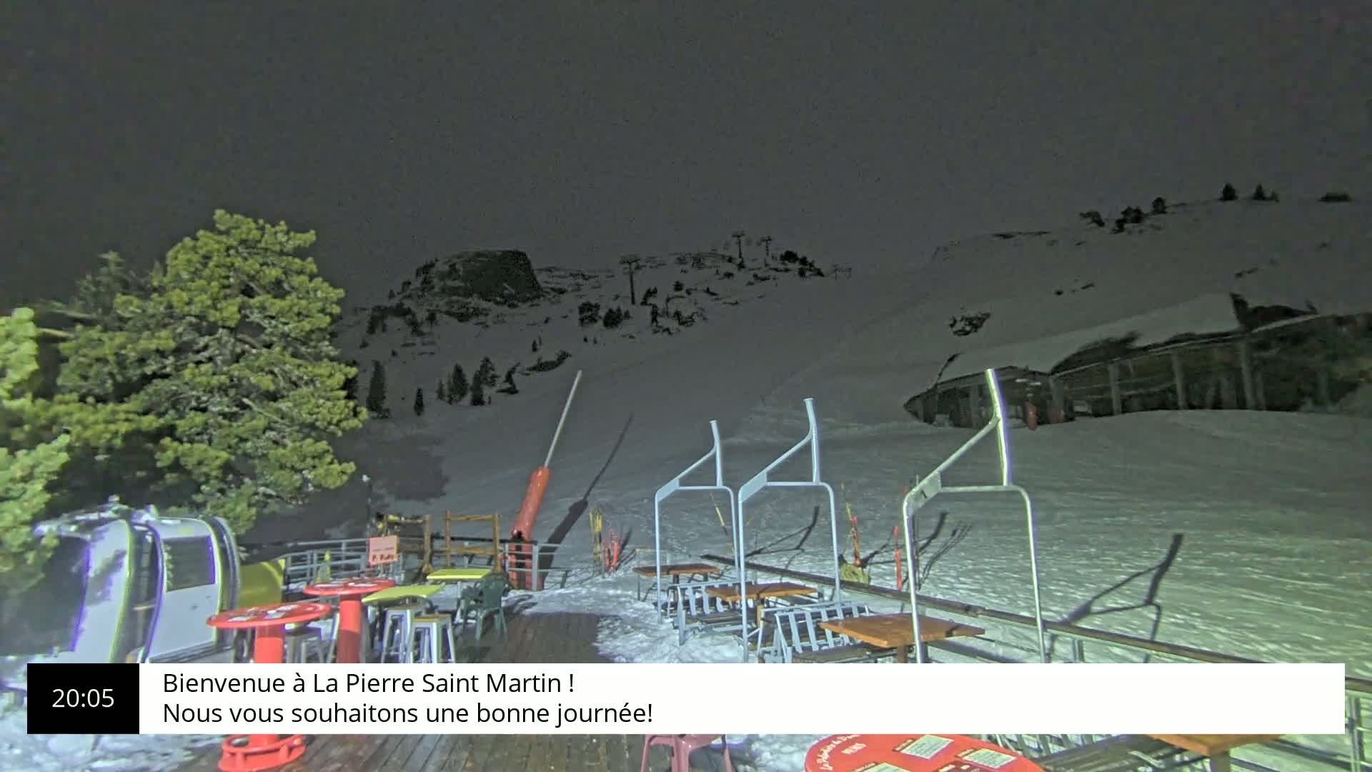 A nighttime view of a snow-covered ski resort with a chairlift, tables, and chairs on a deck, under a dark sky.