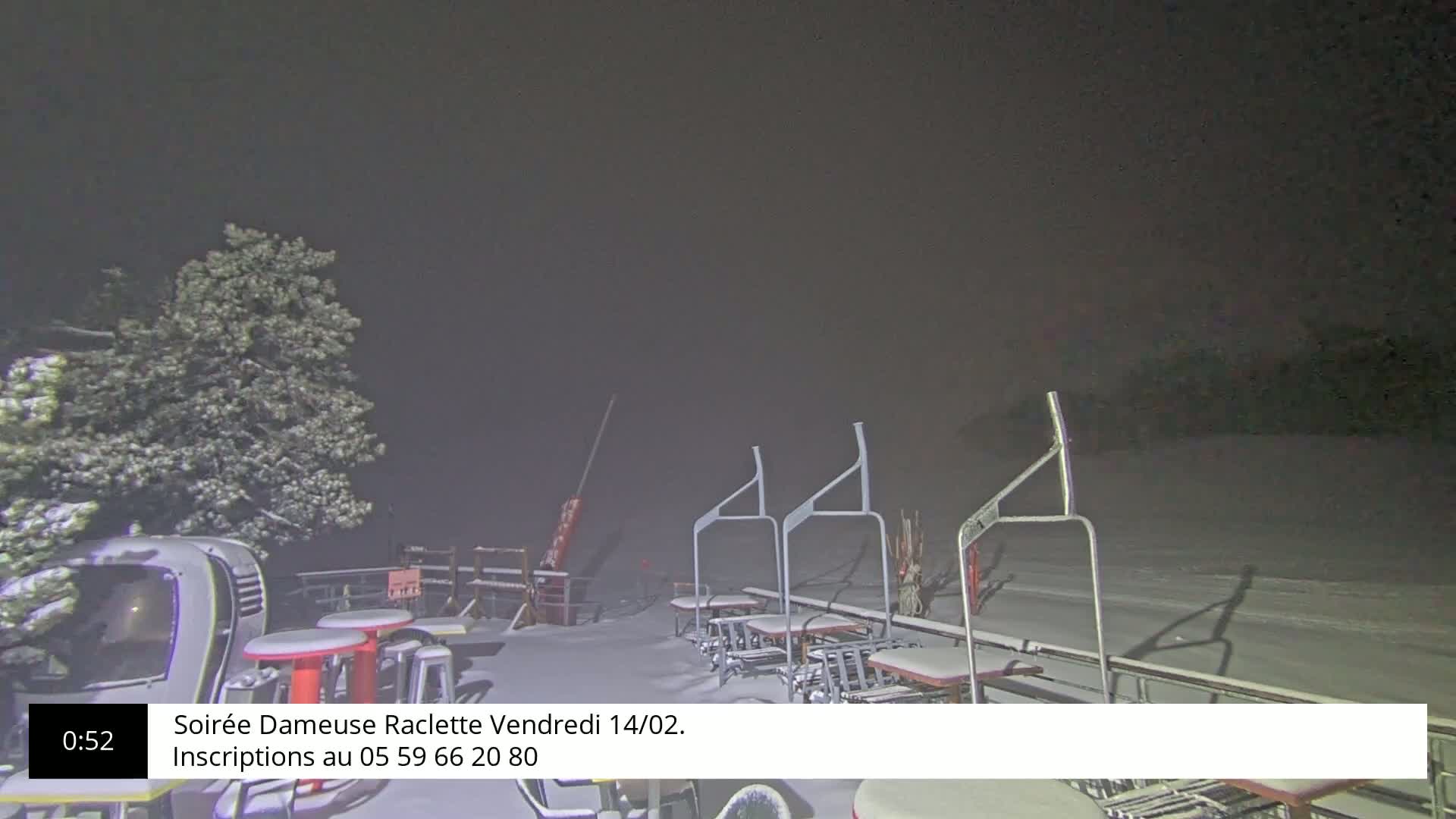 A snowy nighttime scene shows a snow-covered outdoor area with several tables and chairs, a ski lift, and a tree.