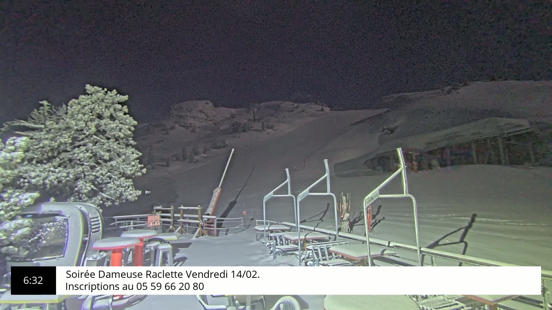 A snow-covered ski slope at night, illuminated by artificial light, shows several empty picnic tables and a chairlift in the foreground, with a snow-laden tree to the left.