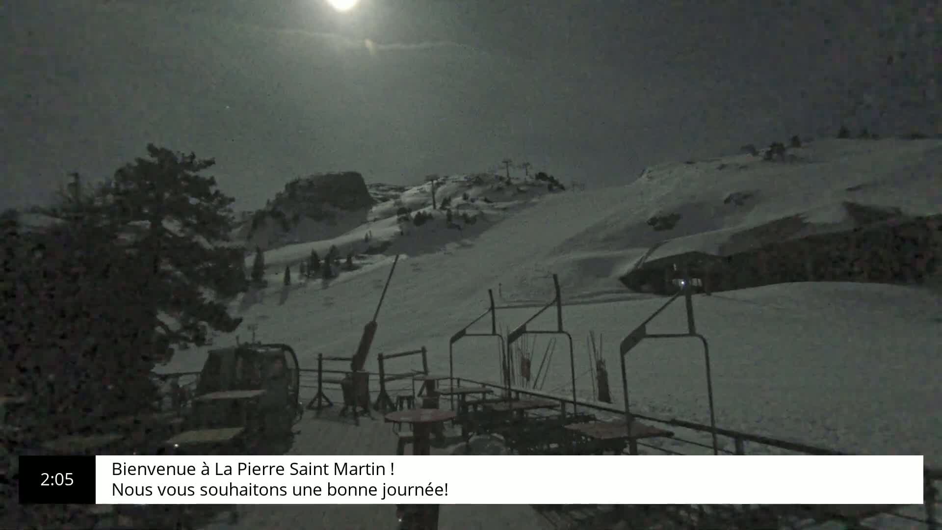A snow-covered ski slope is illuminated by a bright moon at night, with a chairlift and some outdoor seating visible in the foreground.