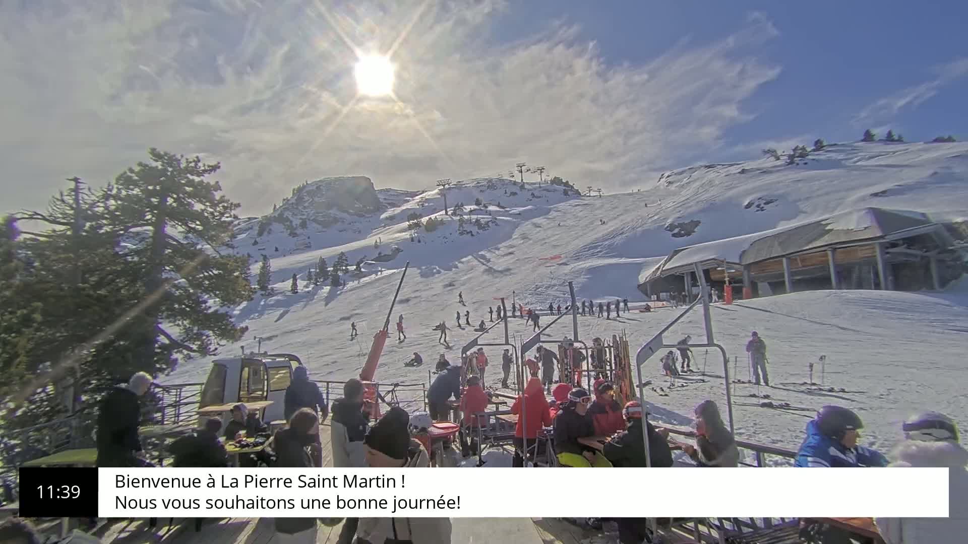 A sunny, snow-covered ski slope with skiers and people relaxing at a base lodge.