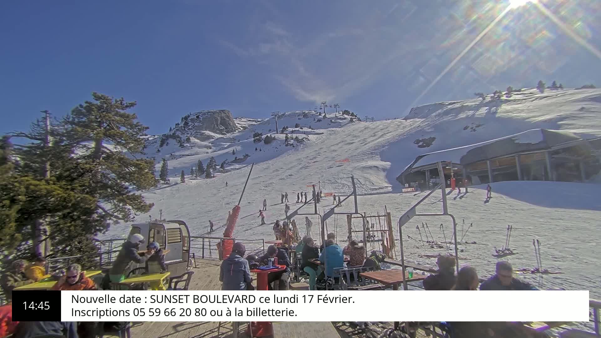 A sunny, snow-covered ski slope with skiers and snowboarders, a ski lift, and a patio area with people sitting at tables.