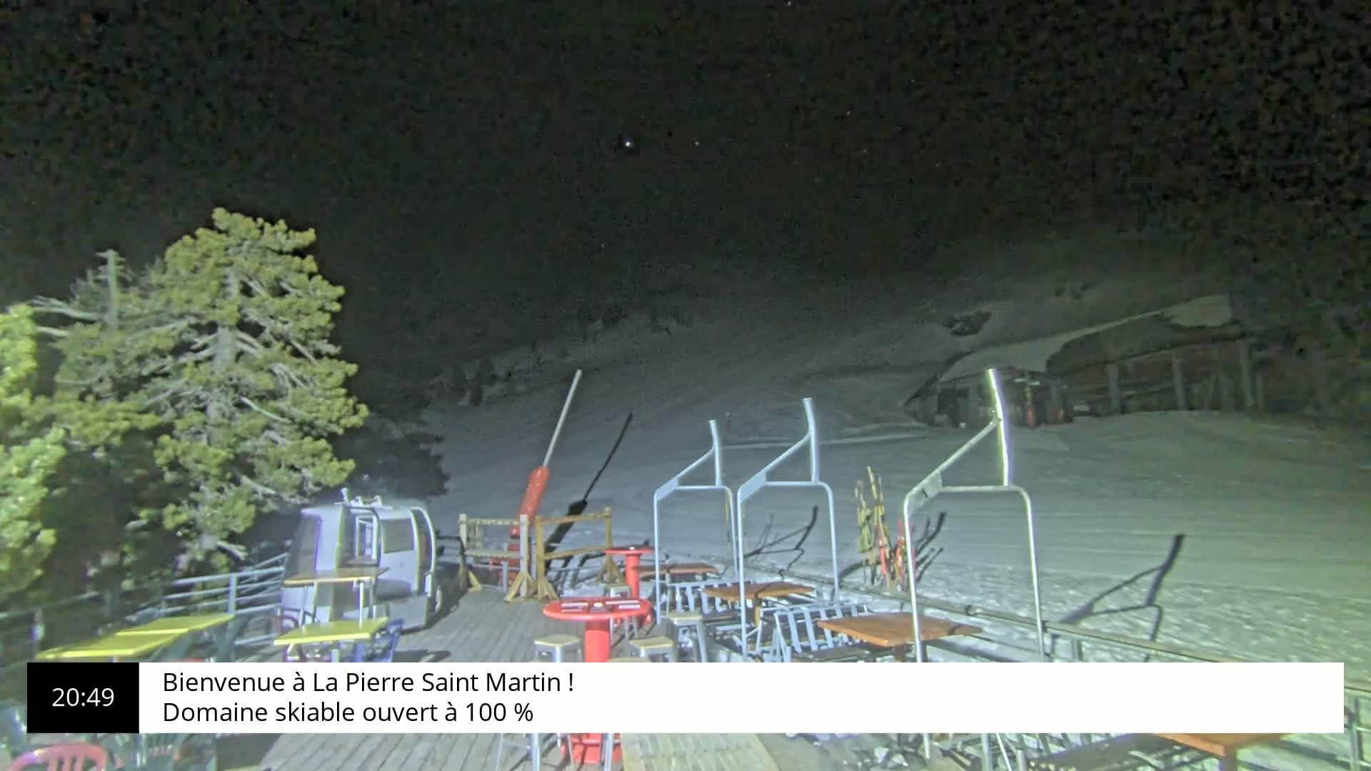 A nighttime view of a snow-covered ski slope with outdoor seating and a chairlift visible under a clear, dark sky.