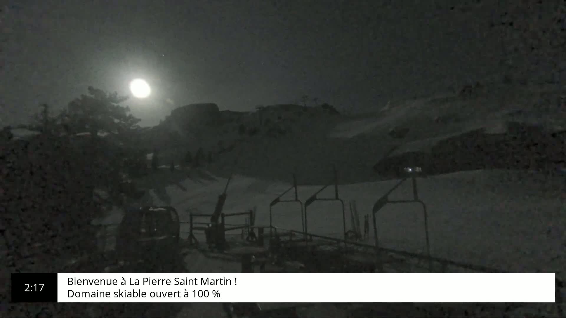 A nighttime view of a snow-covered ski slope illuminated by a bright moon, showing ski lifts and trees in the foreground.