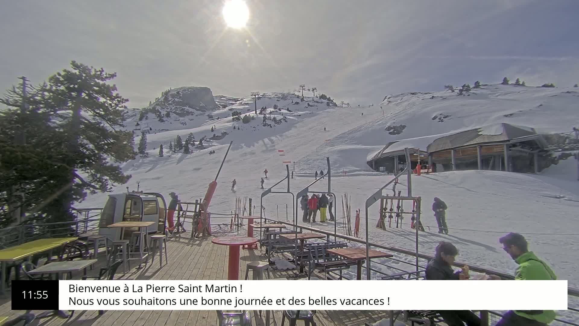 A sunny, snow-covered ski slope with a ski lift, a mountaintop building, and outdoor seating on a deck.