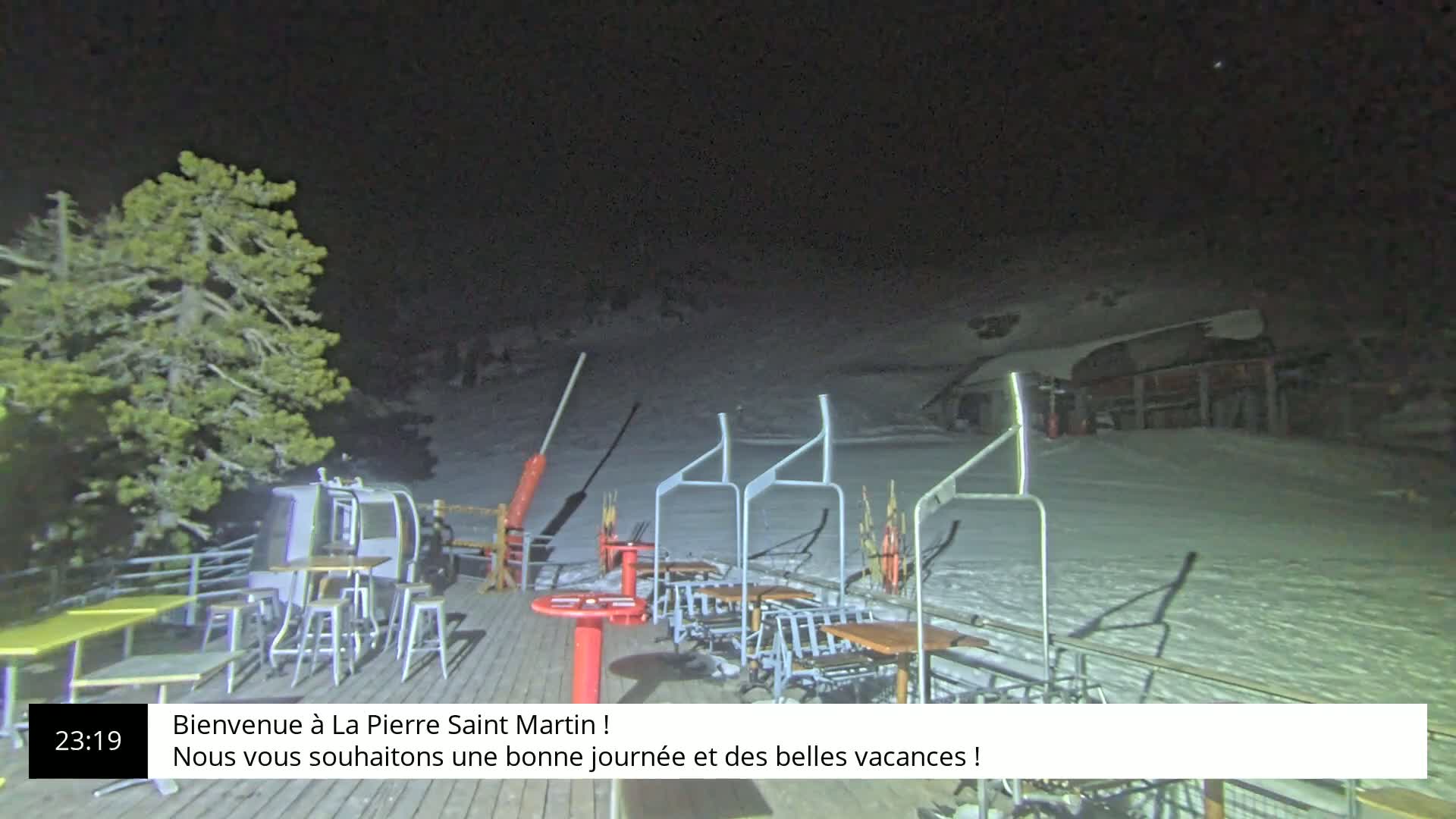 A nighttime view of an outdoor ski lift area with snow-covered ground, several empty tables and chairs, and a tree to the left.