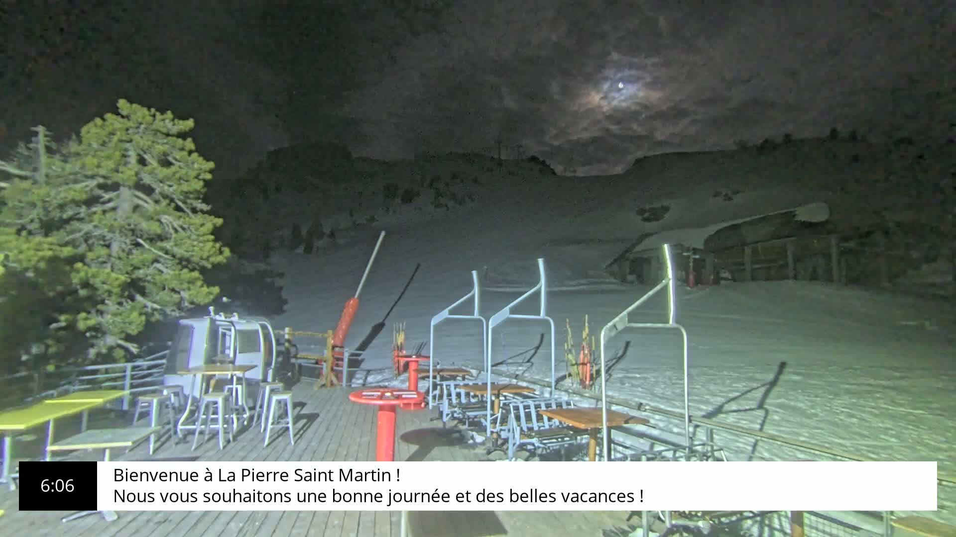 A nighttime scene of a snow-covered ski slope with a moon visible through the clouds, showing outdoor seating and a ski lift.