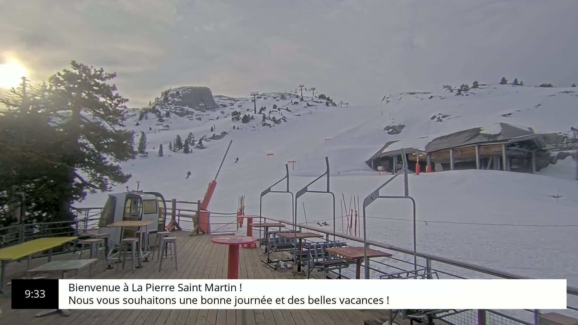 A snow-covered ski slope is visible from a deck with outdoor seating, under an overcast sky.