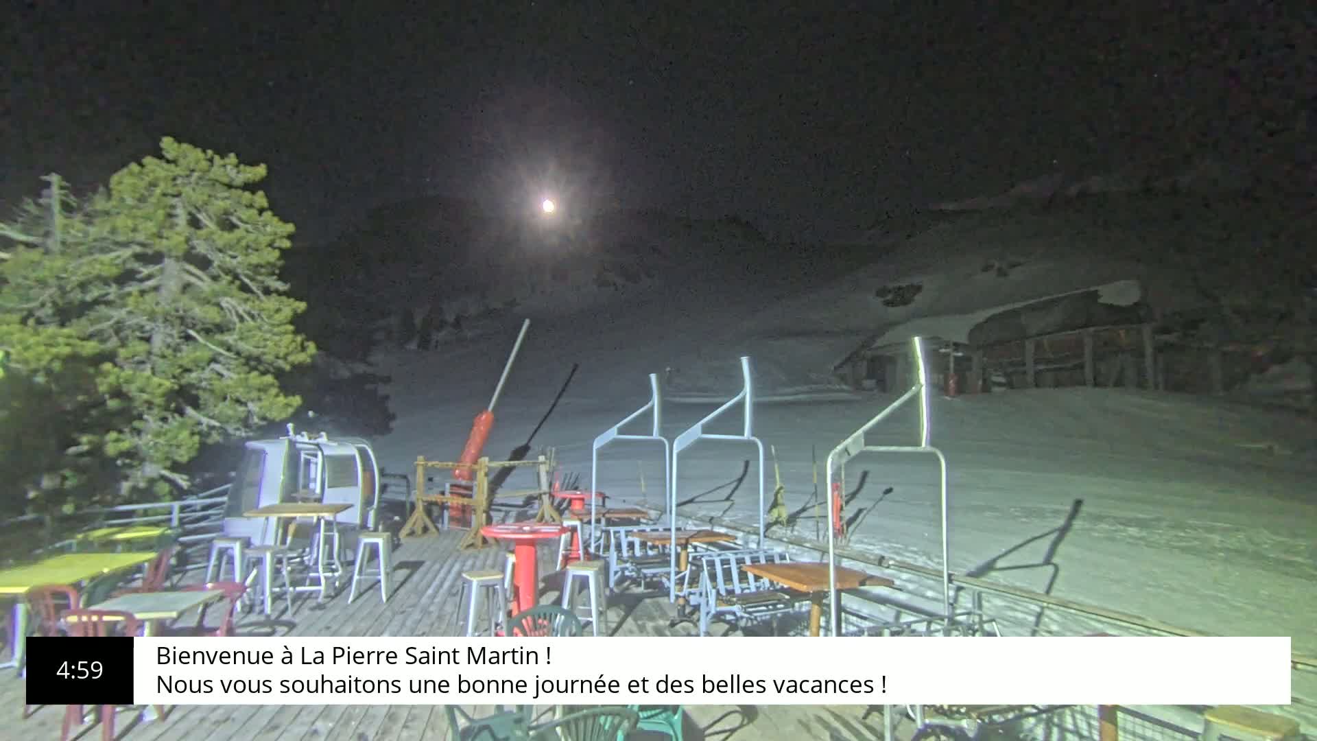 An outdoor nighttime scene shows a snow-covered ski slope with a moonlit sky, outdoor seating and a chairlift.