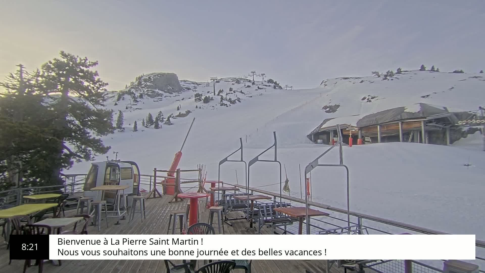 A snow-covered ski slope with a mountain in the background is viewed from a deck with outdoor seating and a chairlift.