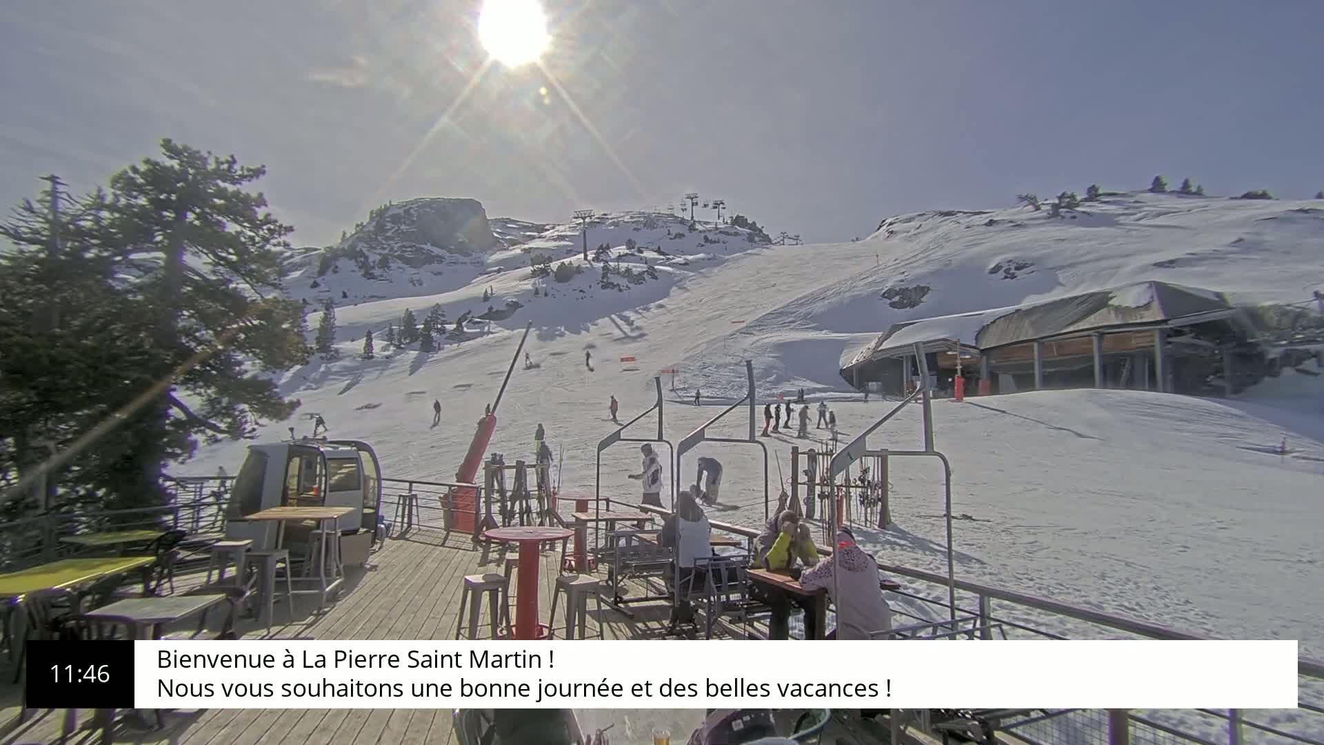 A sunny, snow-covered ski slope with a chairlift, skiers, and a mountaintop restaurant.