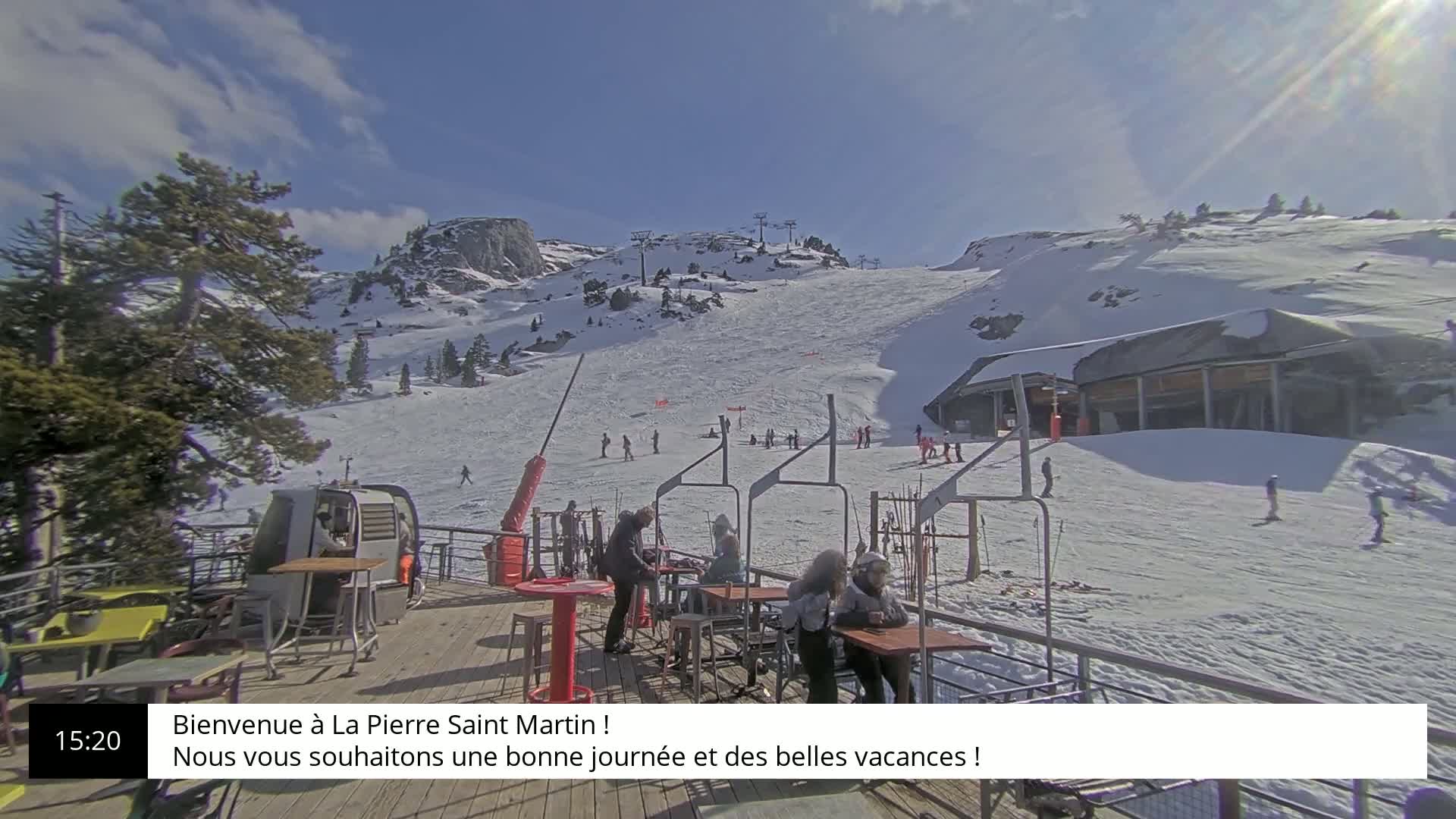 A sunny, snow-covered ski slope with several skiers and snowboarders, a ski lift, and a mountaintop building in the background.