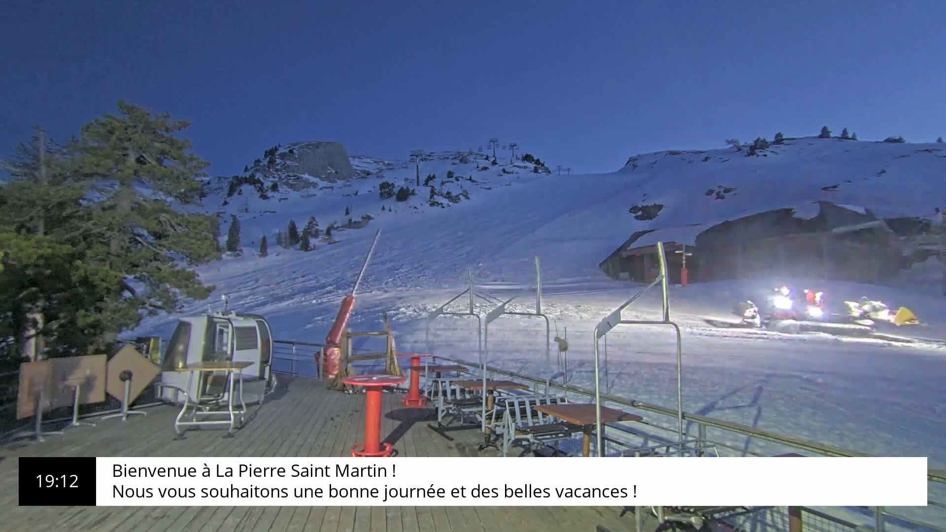 A snow-covered ski slope at twilight, with a chairlift and outdoor seating area in the foreground.